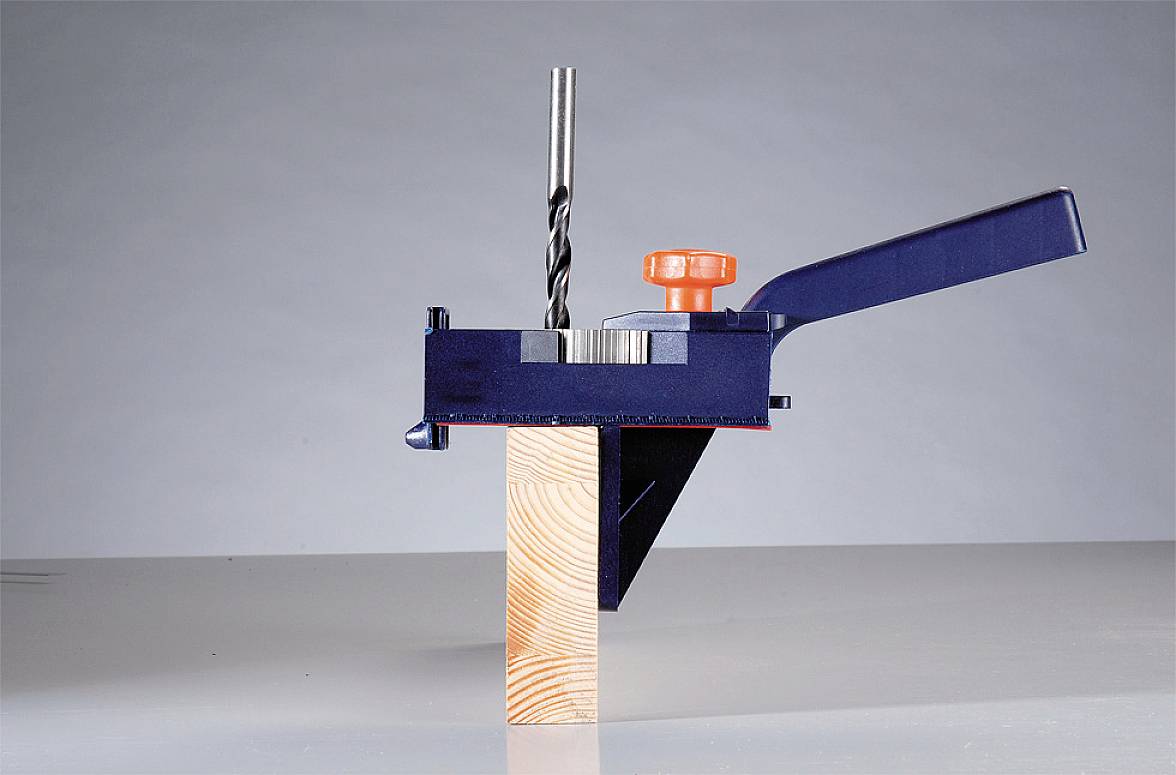 En borr i ett blått fäste borrar ett hål i en träblock på ett bord. Ett orangefärgat handtag är synligt.