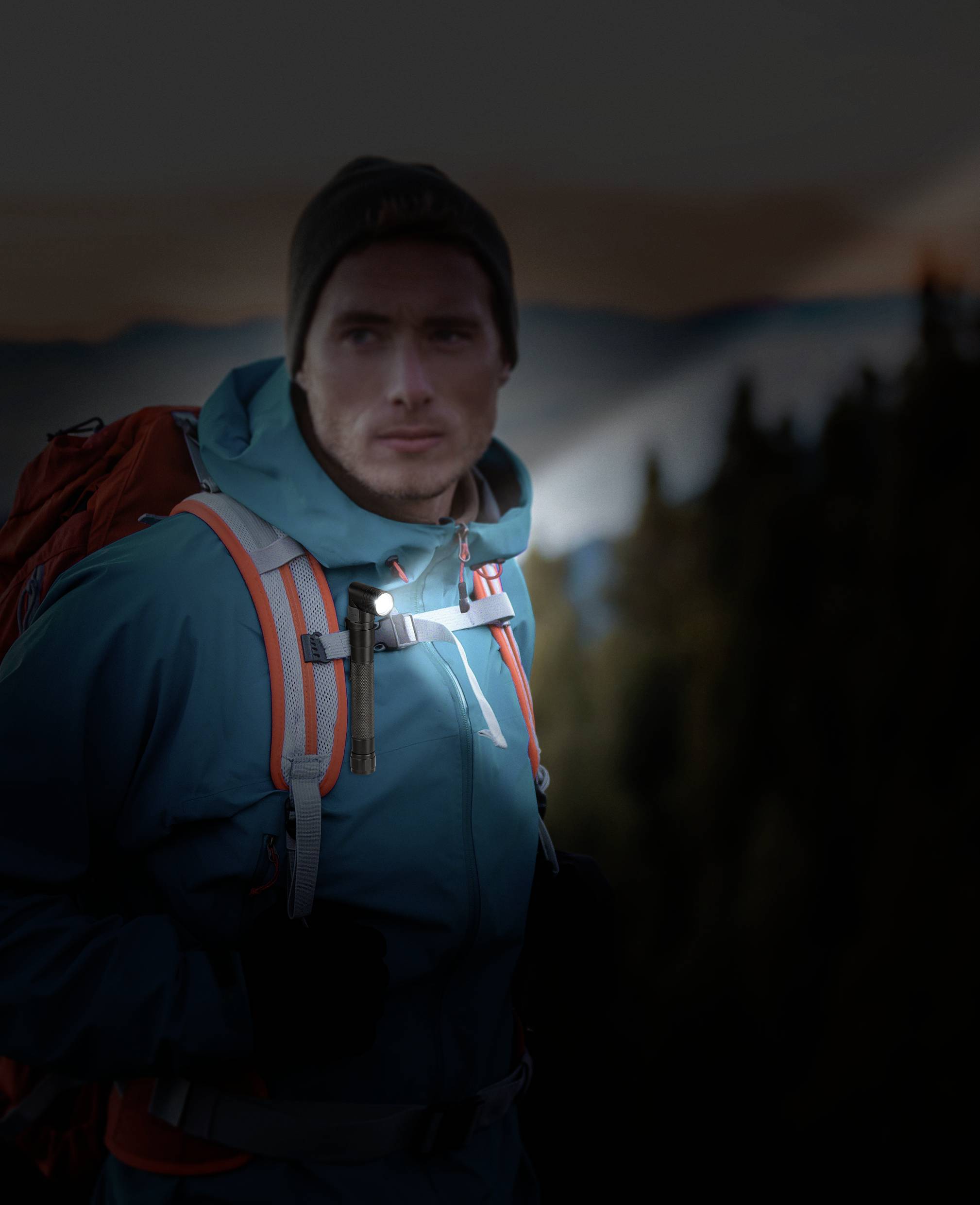 En man i friluftskläder med ryggsäck står i ett bergigt landskap vid skymning. Han blickar uppmärksamt ut över fjärran.
