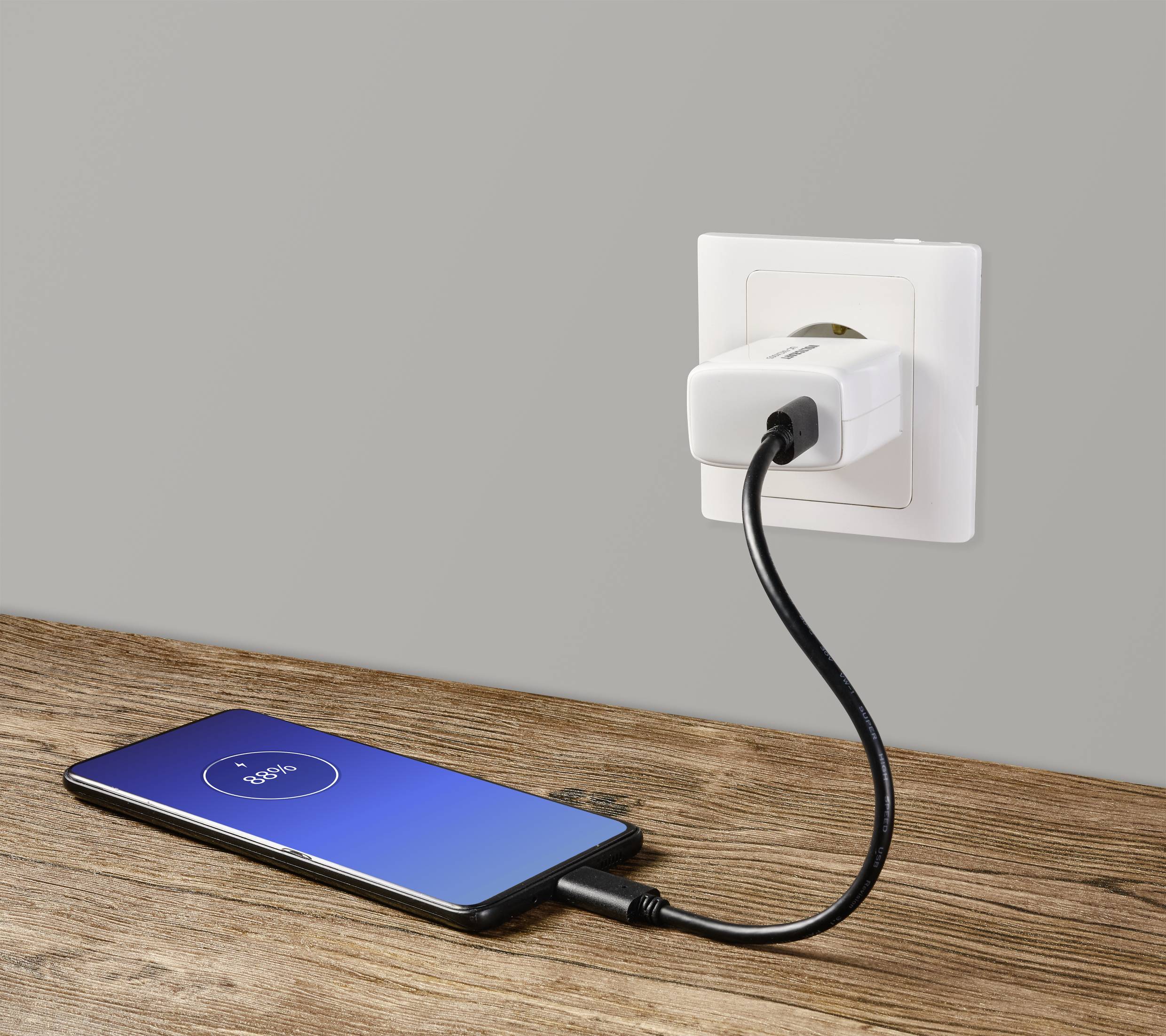 En smartphone ligger på ett träbord och laddas, ansluten med en kabel till ett vitt eluttag i en grå vägg.
