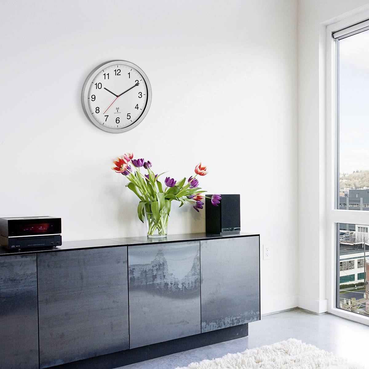 En modern inredning med en svart byrå, på vilken en blombukett står i en vas, och en röd klocka på väggen.