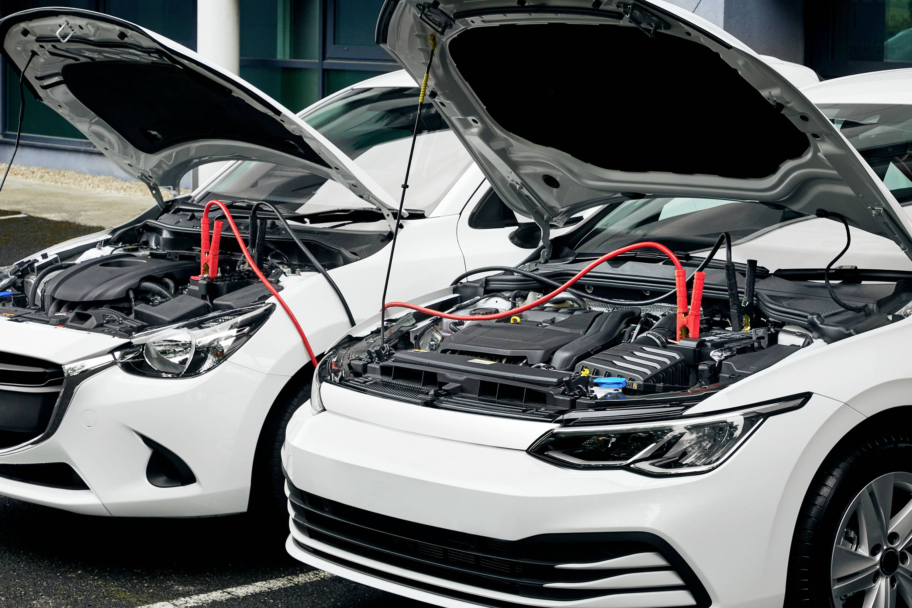Två vita bilar med öppnade motorhuvar står bredvid varandra, sammanlänkade genom röda startkablar som går från batterierna.