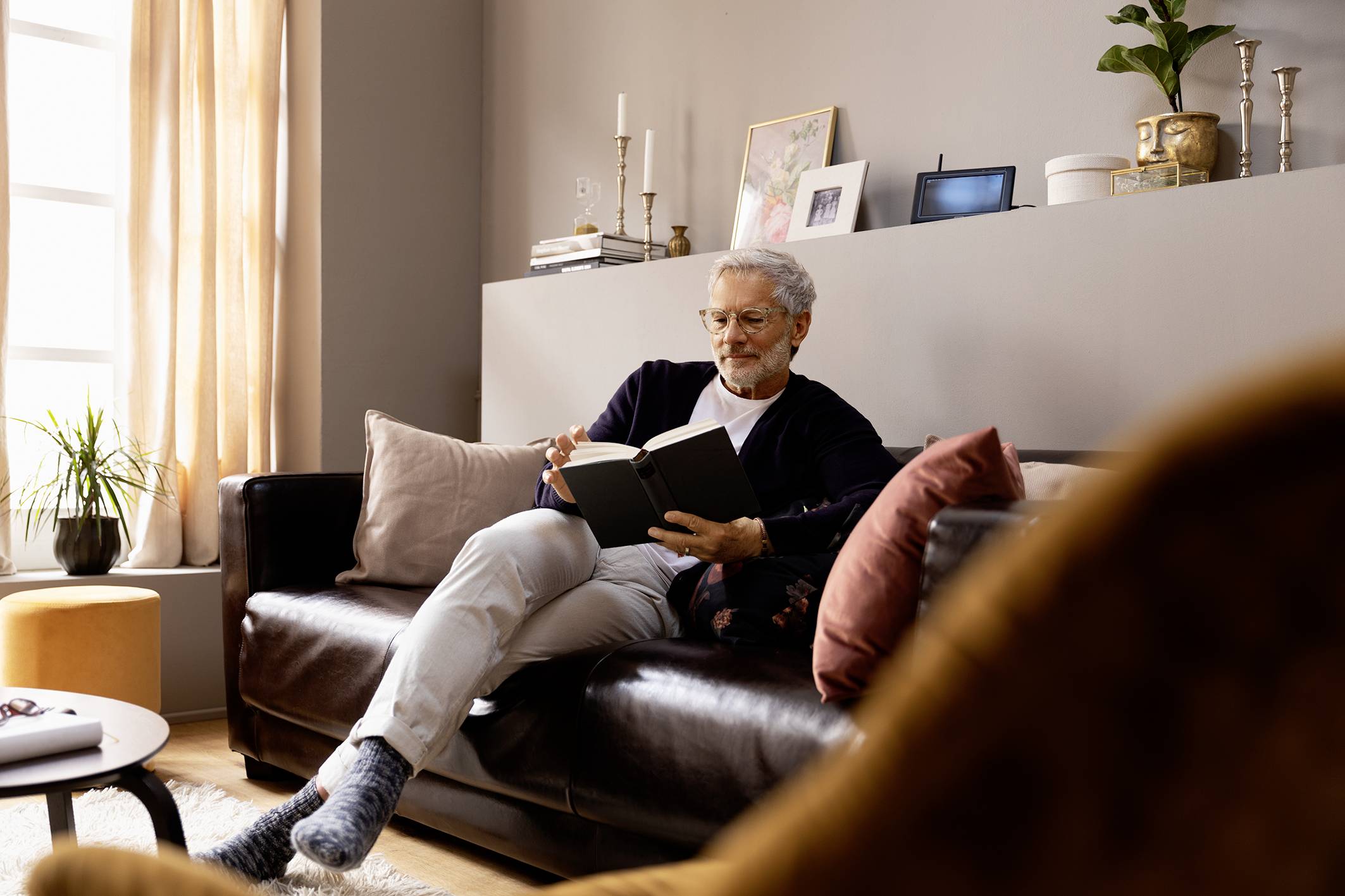 En äldre man sitter avslappnat på en soffa och läser en bok. I bakgrunden syns hyllor med dekorationer och växter.