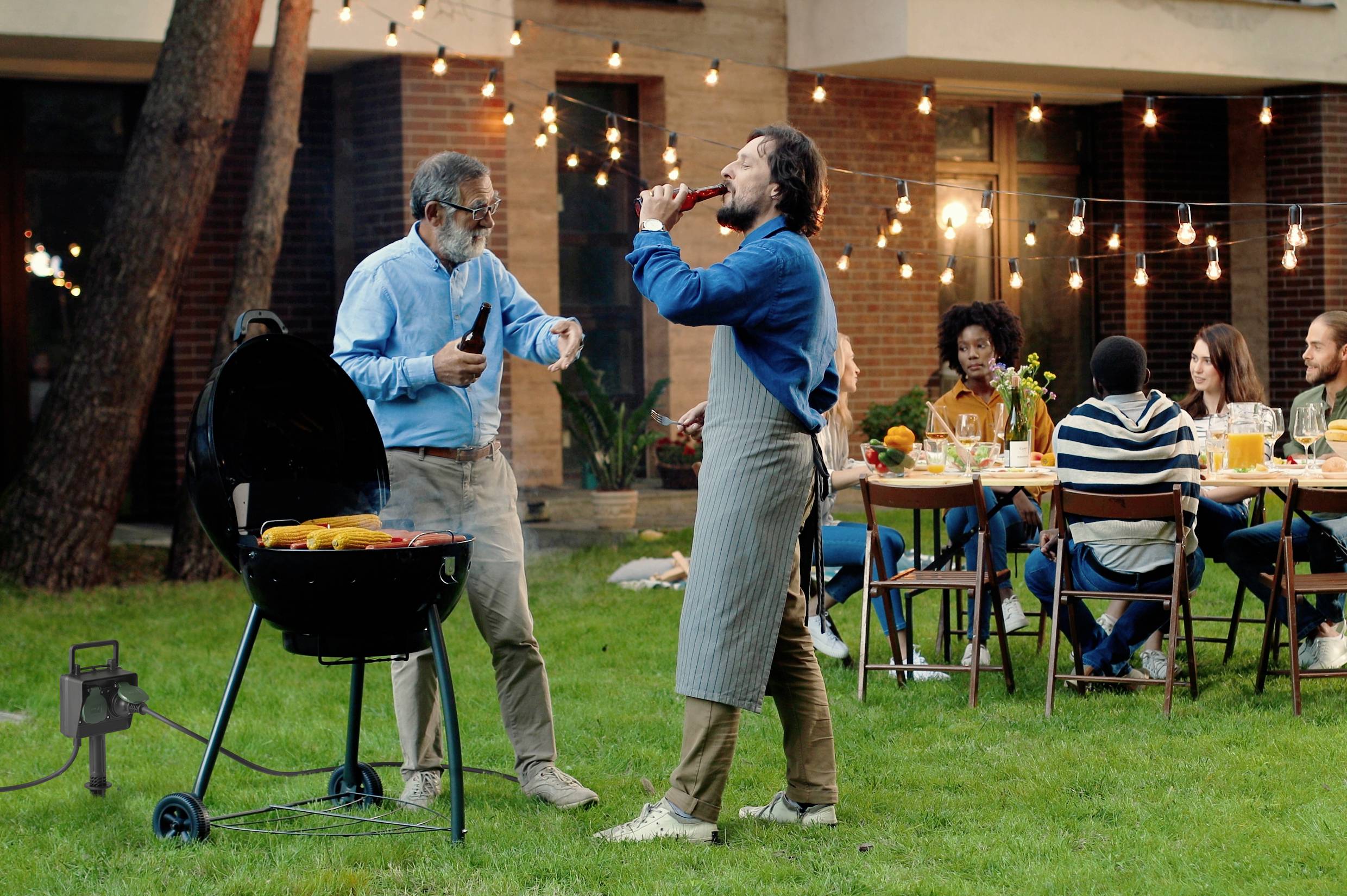 En grupp människor sitter vid ett dukat bord i trädgården. Två män står vid grillen, medan en annan njuter av en drink. Lampor är upphängda.