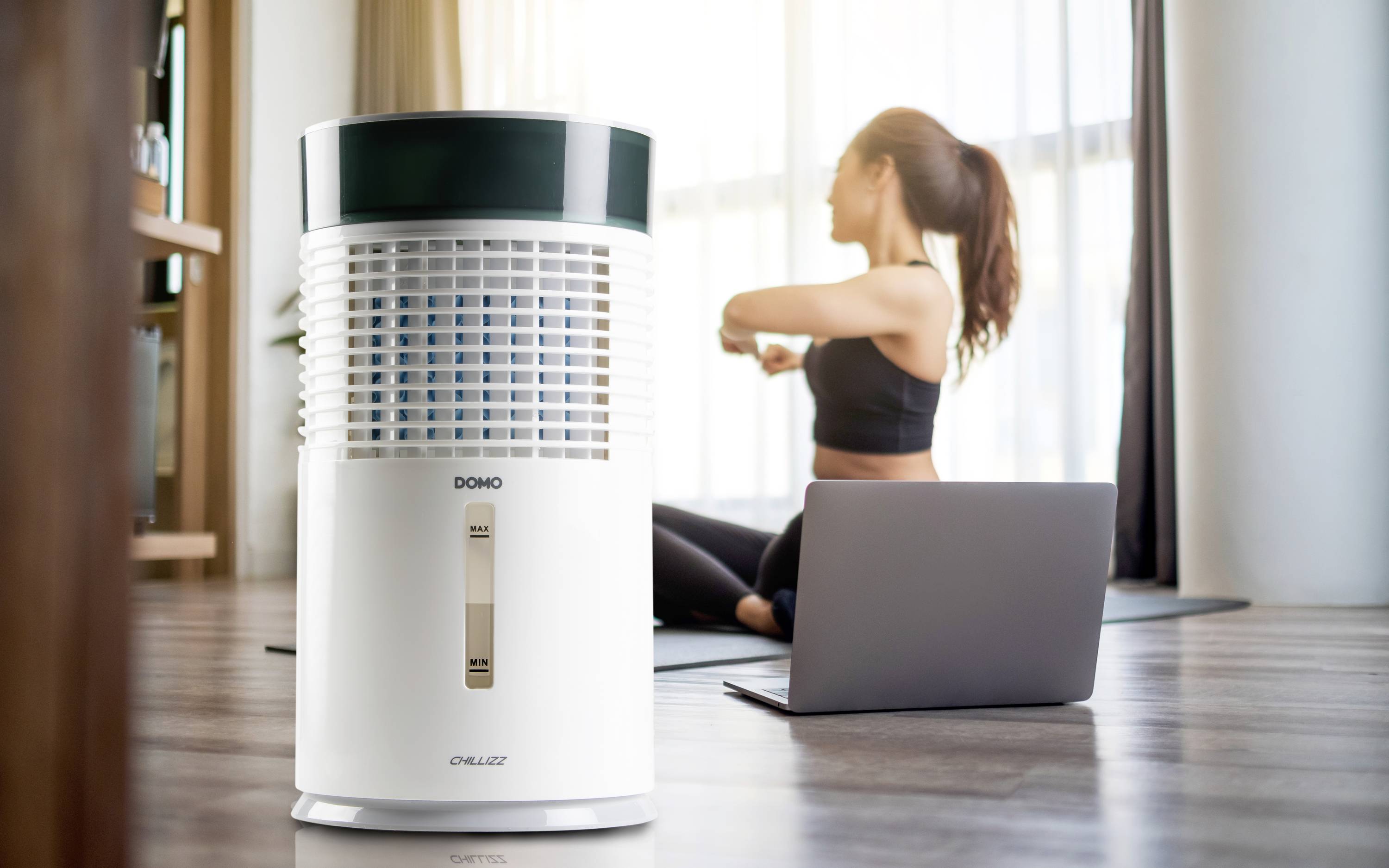 Luftrenare i förgrunden, kvinna gör yoga i bakgrunden på en yogamatta med en laptop bredvid. Ljus rumslig omgivning med ett fönster.