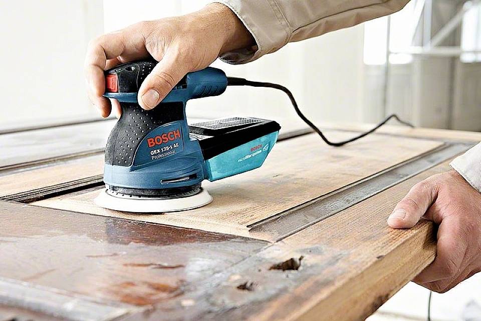 En person slipar en träyta med en elektrisk oscillerande slipmaskin. Sliparen är av märket Bosch.