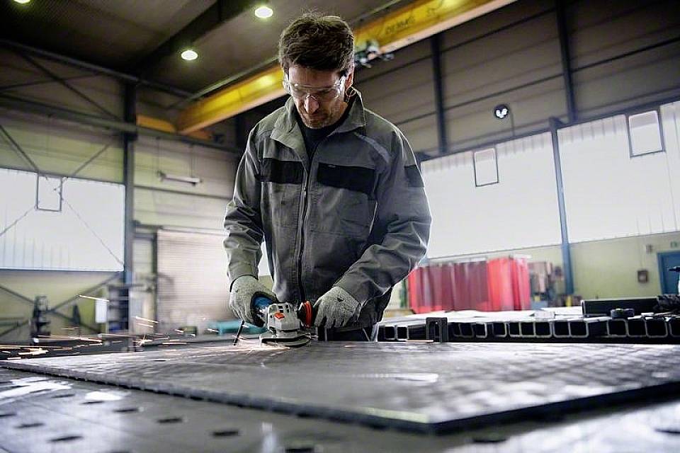 En arbetare i en verkstad slipar en metallplatta med ett elverktyg. Gnistor flyger, omgivningen är industriell.