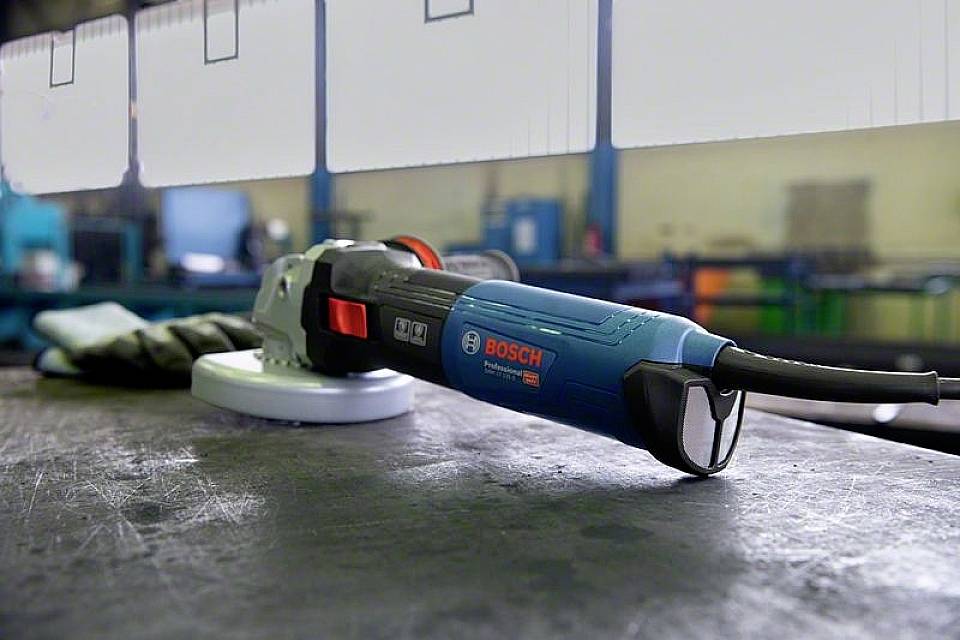En blå vinkelslip från Bosch ligger på ett bord i en verkstad. I bakgrunden syns oskarpa maskiner.