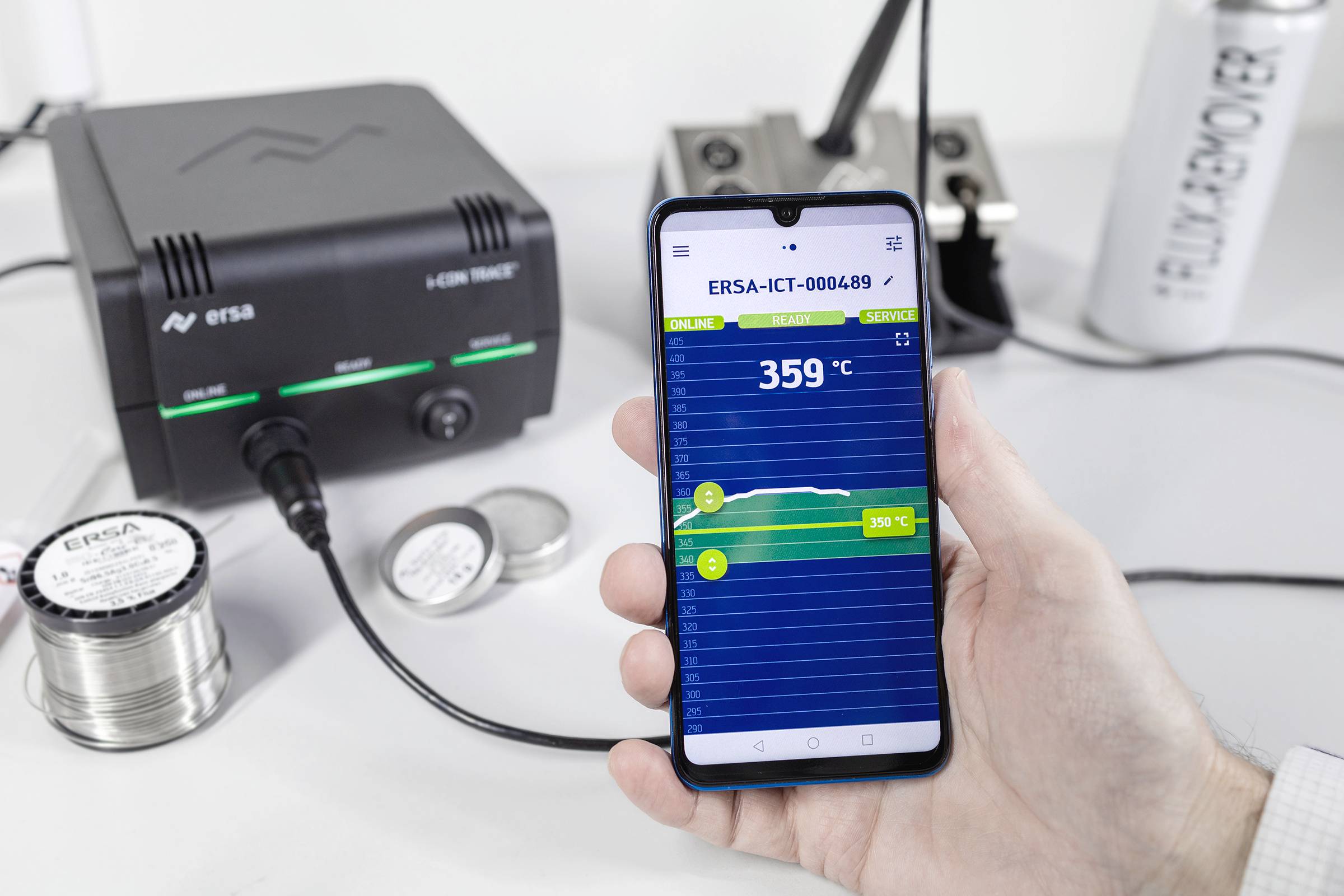 En hand håller en smartphone med en temperaturvisning på 359 °C. I bakgrunden syns en lödstation med lödtennrullar och ett varumärkesnamn.