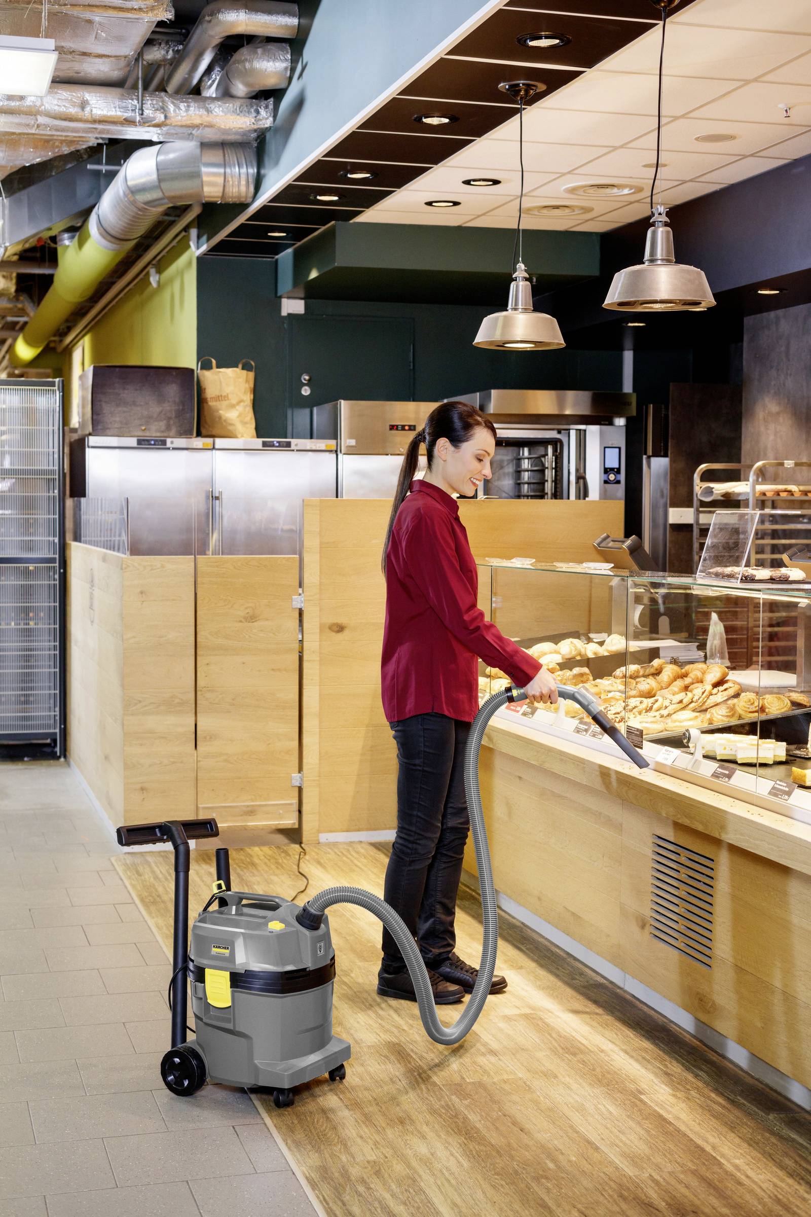 En kvinna i en röd tröja dammsuger ett bageris golv nära en display med bakverk. Industriella takrör och träpanel syns.