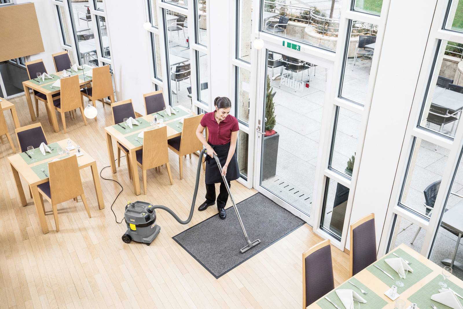 En person dammsuger en dörrmatta vid ingången till en ljus, tom restaurang. Bord är dukade med bordstabletter och servetter. Stora fönster erbjuder utsikt utomhus.