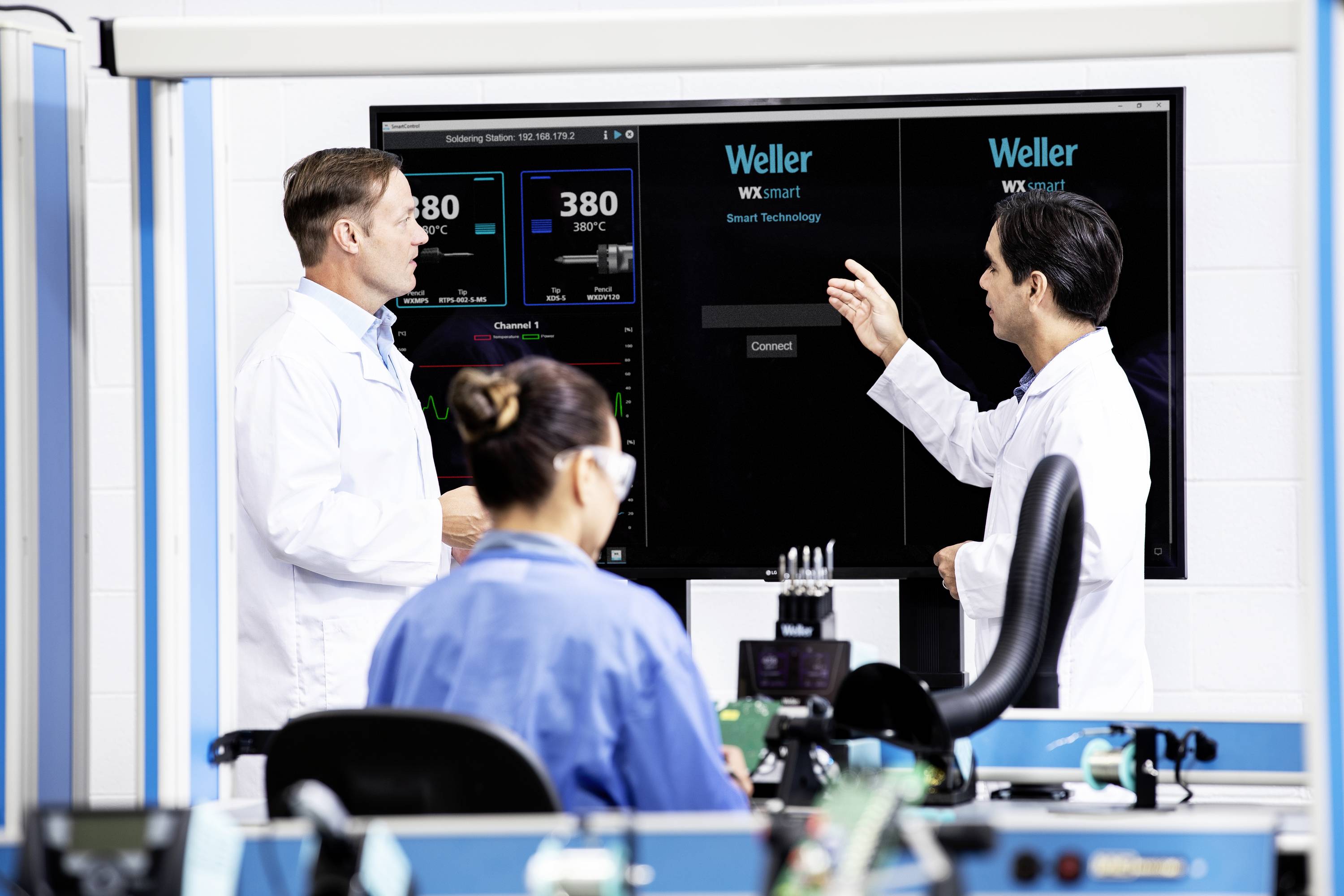 Två män och en kvinna i laboratoriekläder arbetar med elektroniska enheter. En av männen pekar på en skärm med tekniska data.