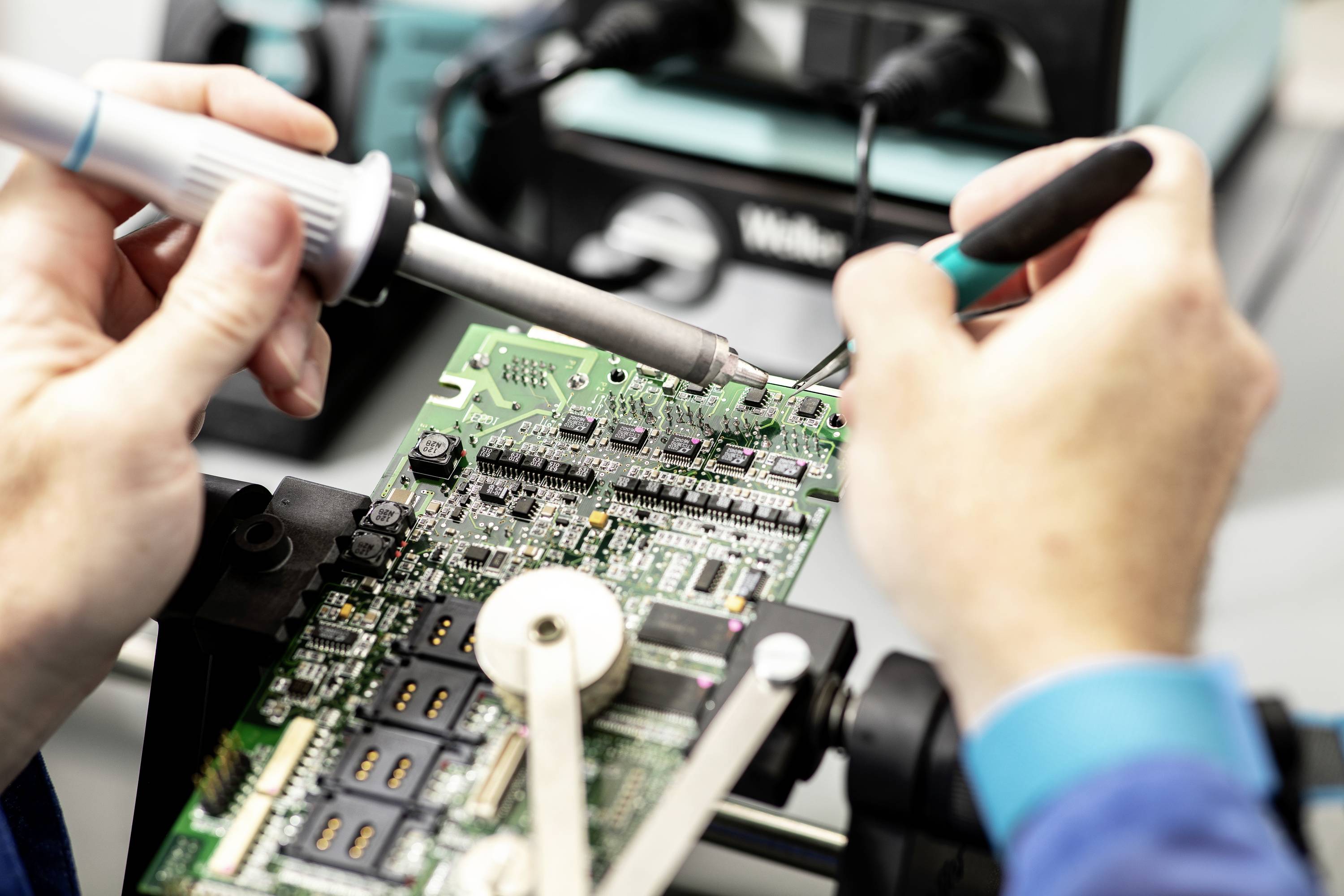 En person löder noggrant på en elektronisk kretskort, omgiven av tekniska enheter, i en laboratoriemiljö.