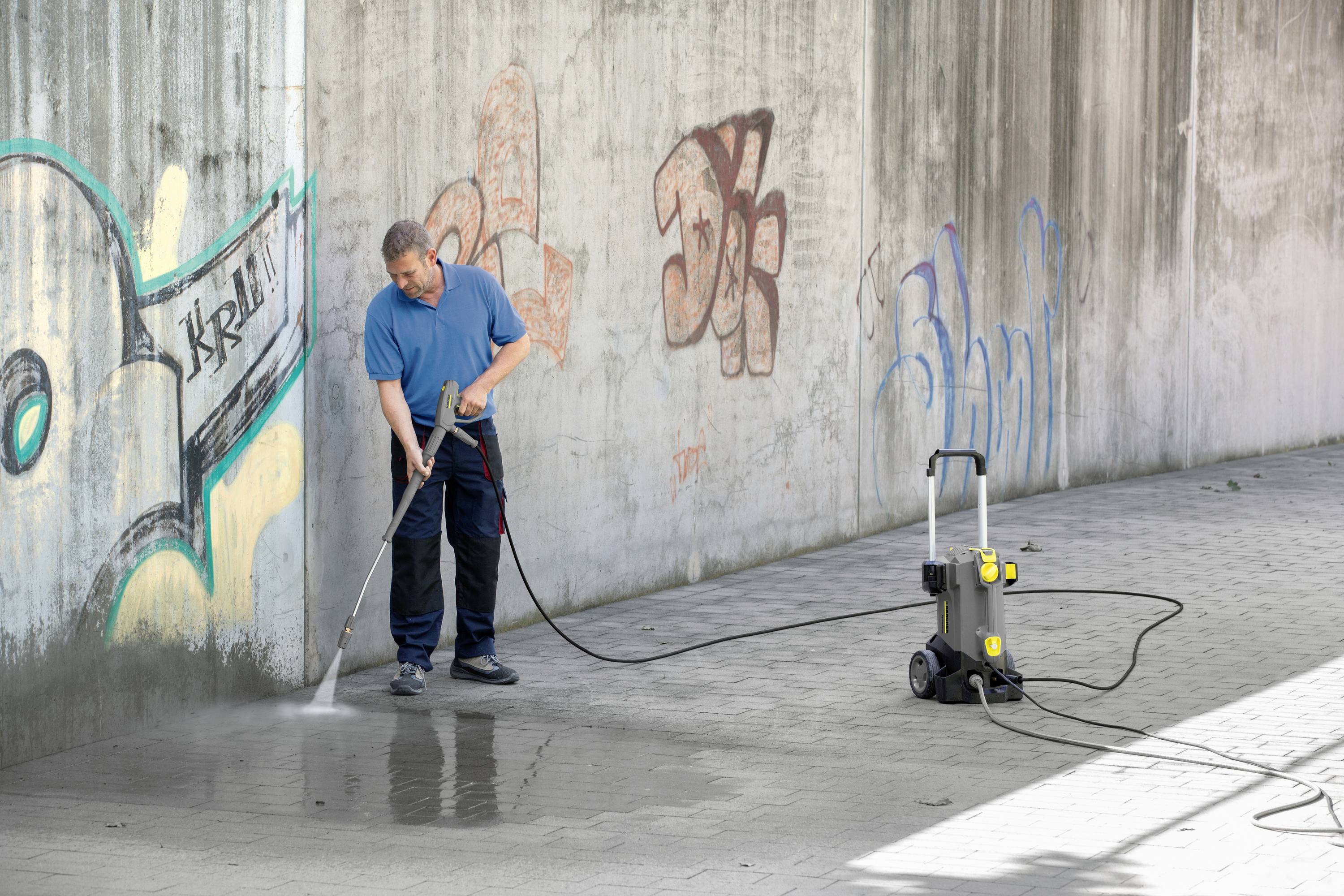 En person rengör en med graffiti täckt mur med en högtryckstvätt på ett stenlagt golv.