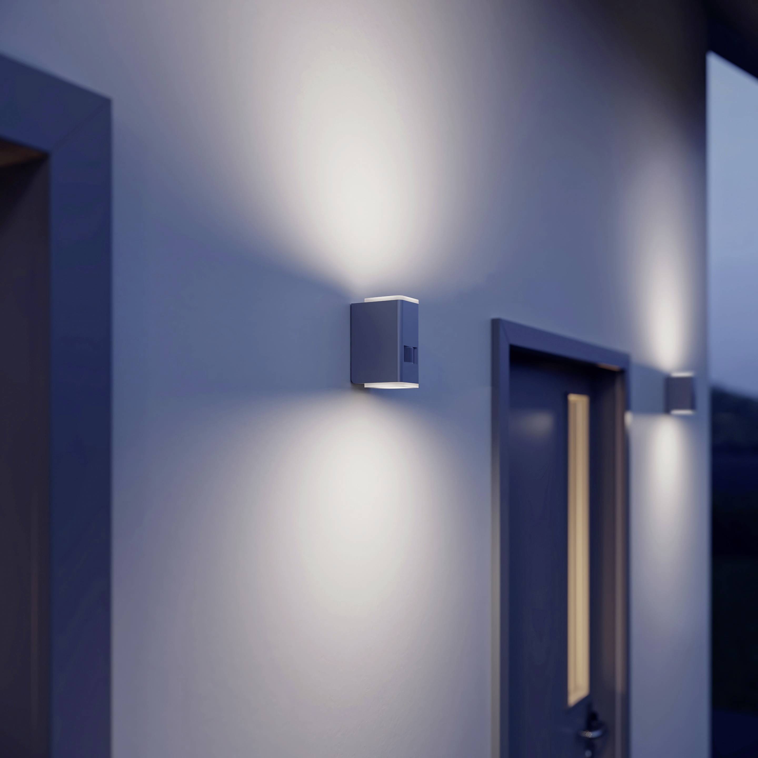 En yttervägg på ett hus vid skymning, belyst av en modern vägglampa som kastar ett mjukt ljus uppåt och nedåt.