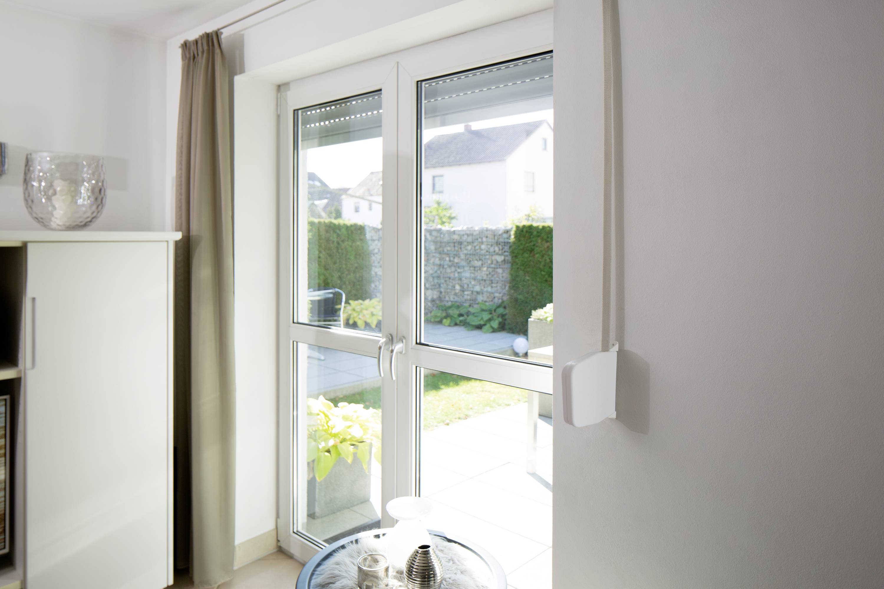 Ett ljust bostadsutrymme med glasdörrar som leder ut till en terrass och därutöver visar en välskött trädgård med häckar och mur.