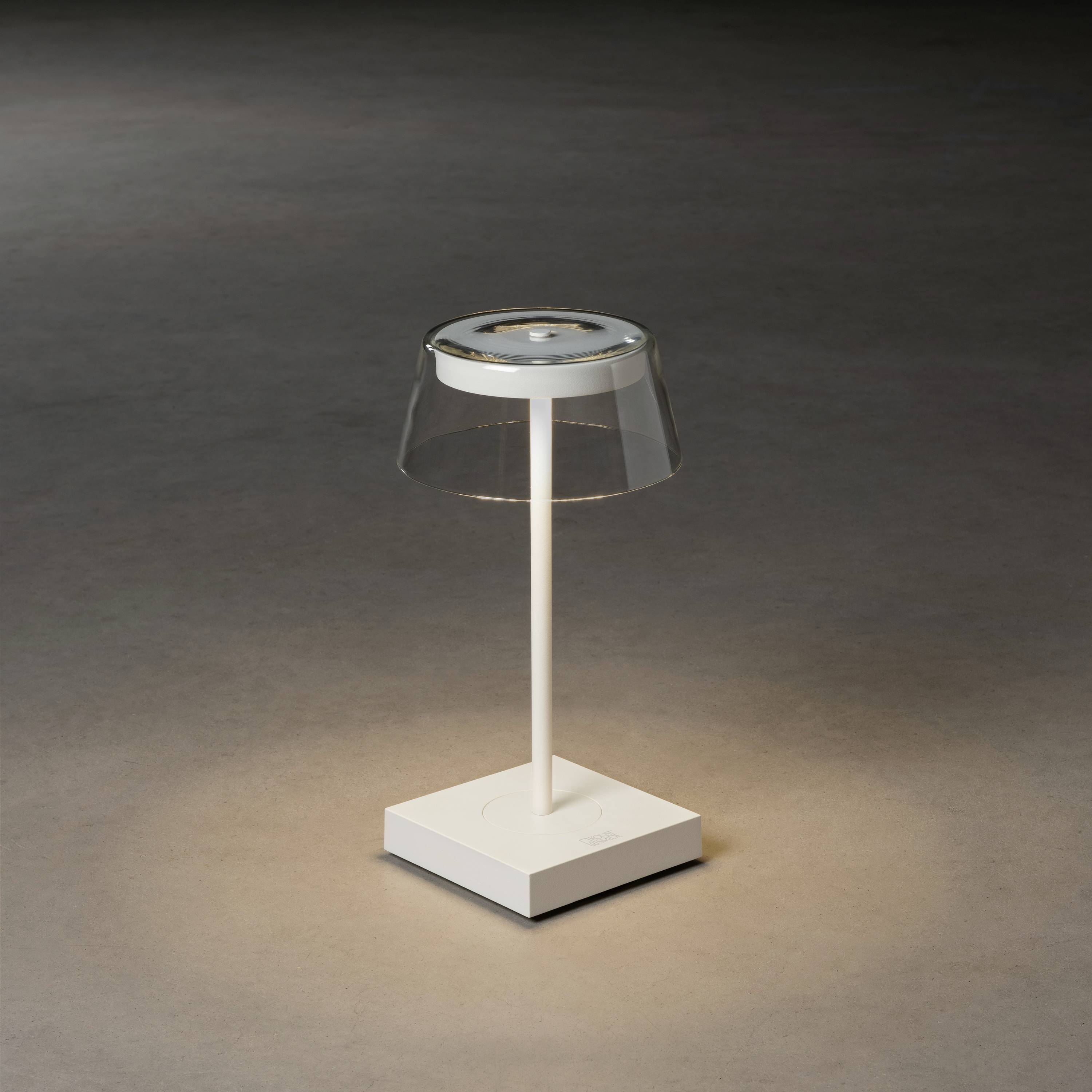 En modern bordslampa med transparent skärm och vit sockel lyser mjukt på en mörk yta.