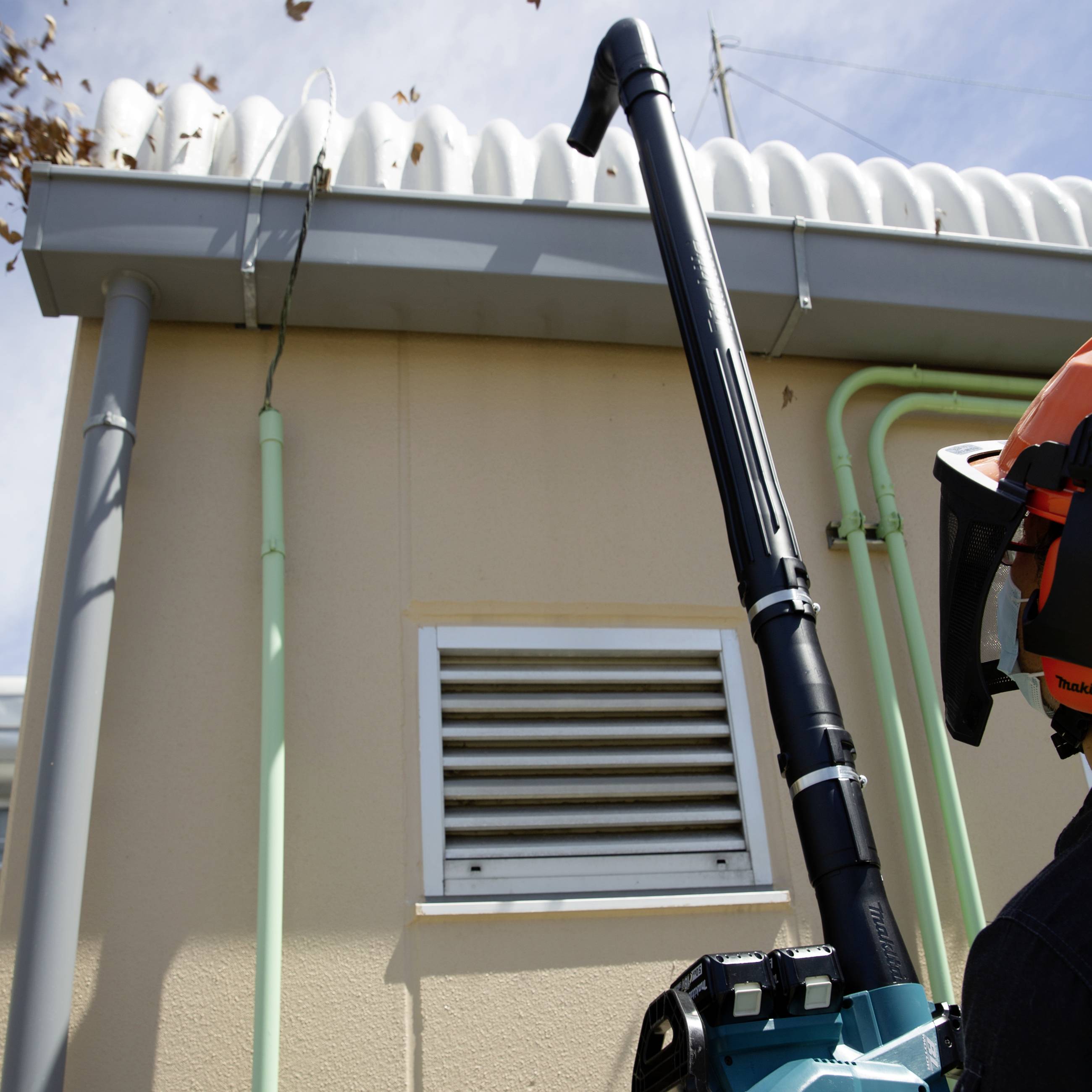 En person använder ett lövblås för att ta bort löv från ett tak. En byggnad och rör syns i bakgrunden.