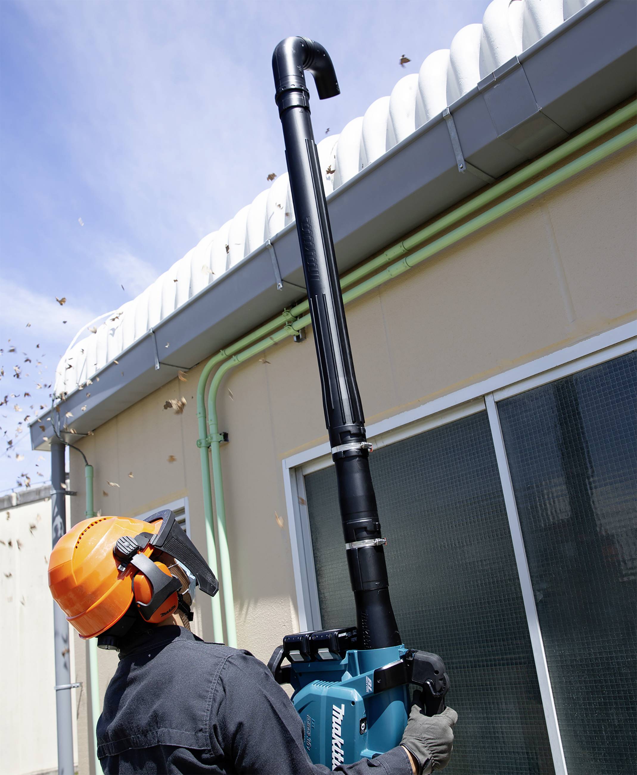 En person står på marken och använder en maskin för att ta bort löv från ett högt tak. Personen bär en skyddshjälm.