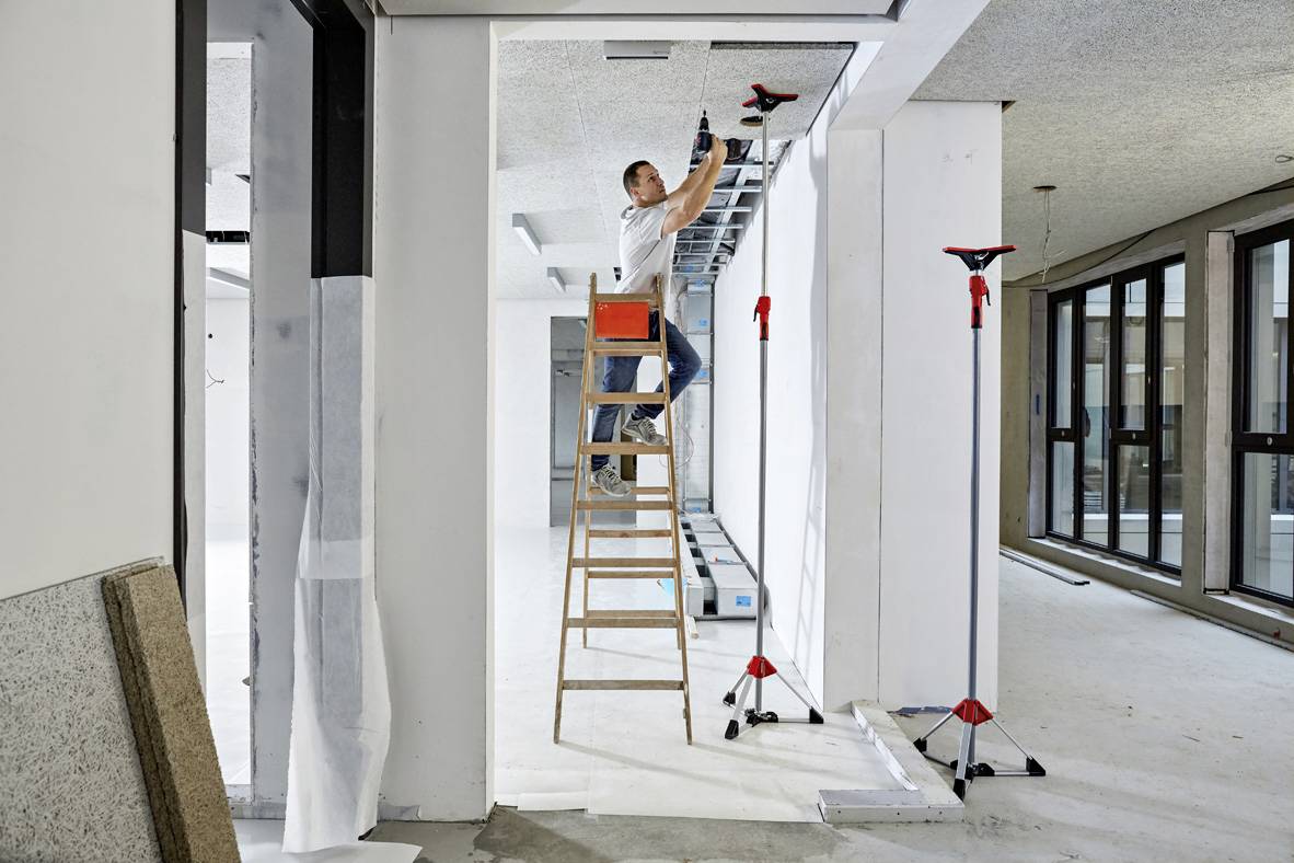 En hantverkare står på en stege och installerar en metallstruktur i taket i ett oavslutat, modernt rum.