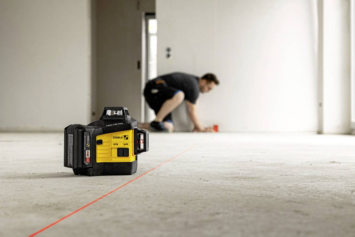 En gul lasernivåinstrument projicerar en röd linje på golvet. I bakgrunden knäböjer en person och arbetar.
