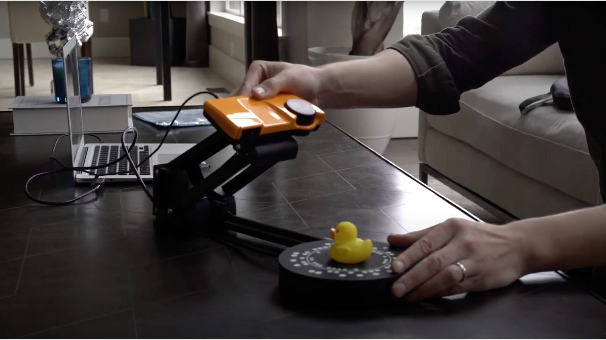 En person skannar en gul gummianka på en rund roterande skiva med en orangefärgad 3D-skanner på ett bord.