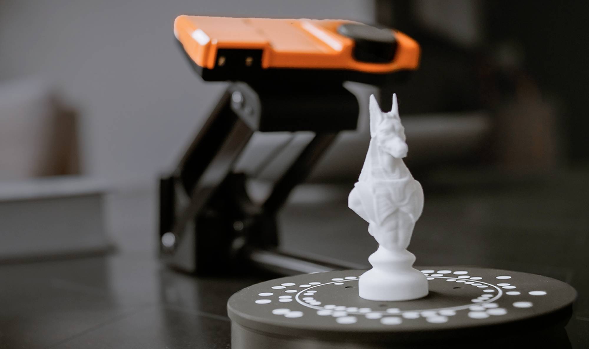 Bilden visar en vit schackspringare på ett runt bord med en 3D-skanner bakom. Skannern är orange och svart.