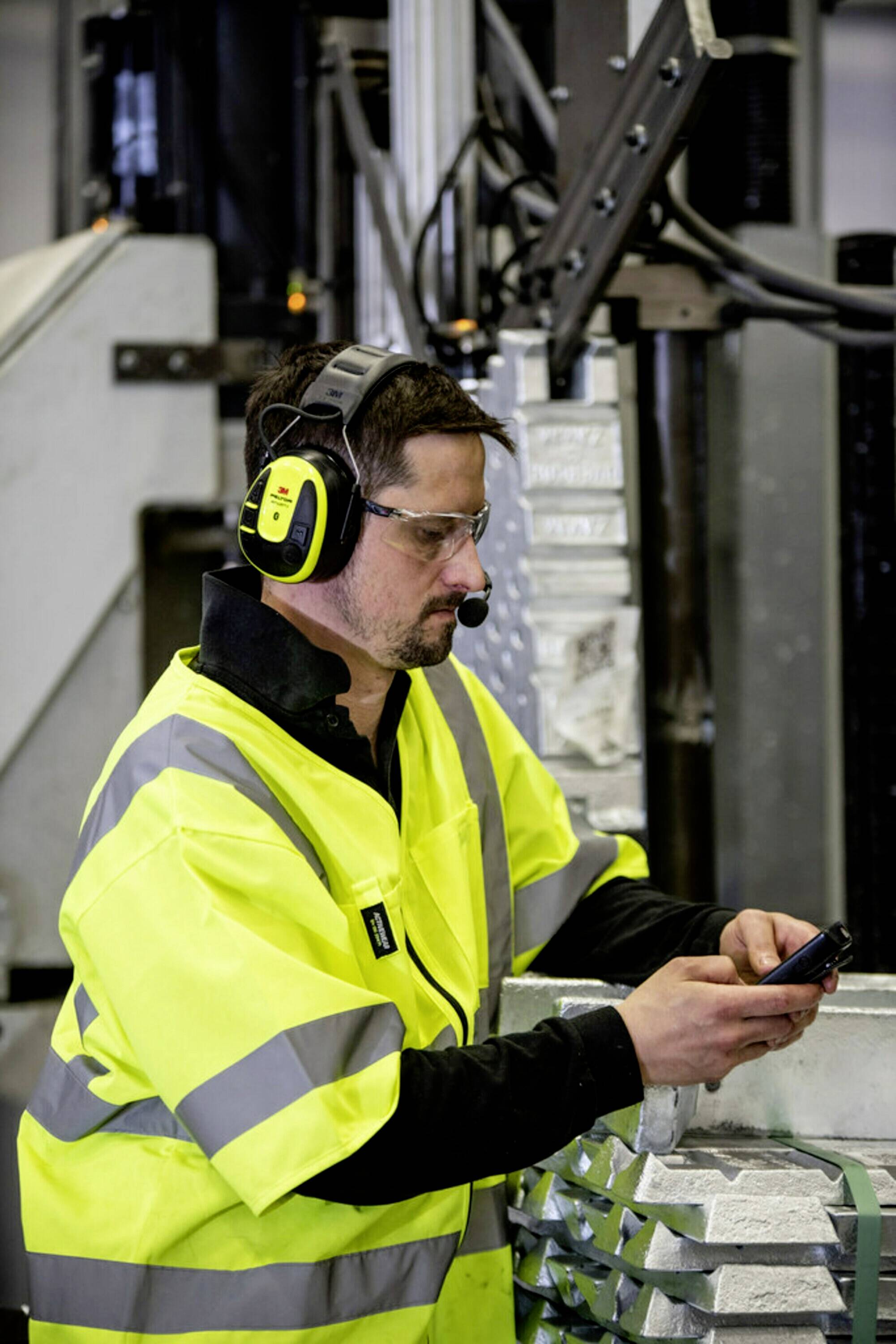 Man i gul varselväst och skyddsglasögon, arbetar vid en enhet i en fabriksmiljö, bär hörlurar med mikrofon.