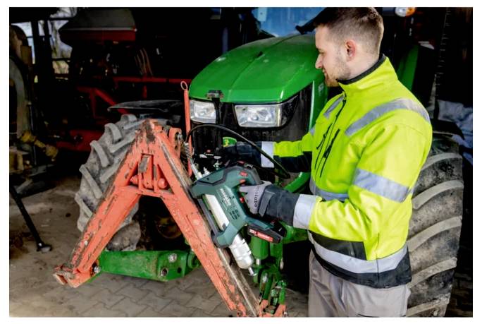 Mekaniker i en reflexväst använder ett elverktyg för att serva frontfästet på en grön traktor i en verkstad.