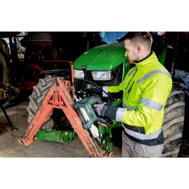 En person i en fluorescerande jacka använder ett elverktyg för att arbeta på en traktor i en verkstadsmiljö.