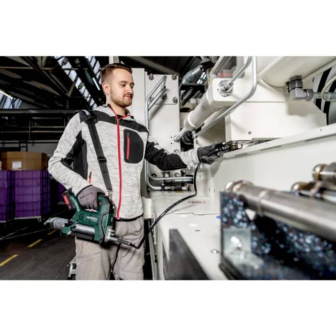 En person som använder en eldrillborrmaskin på industriell maskineri i en fabriksmiljö, omgiven av utrustning och lagringsutrymmen.