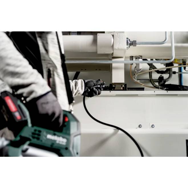 En person i arbetshanskar håller ett elverktyg och justerar maskineri i en industriell miljö med synliga rör och metalliska ytor.