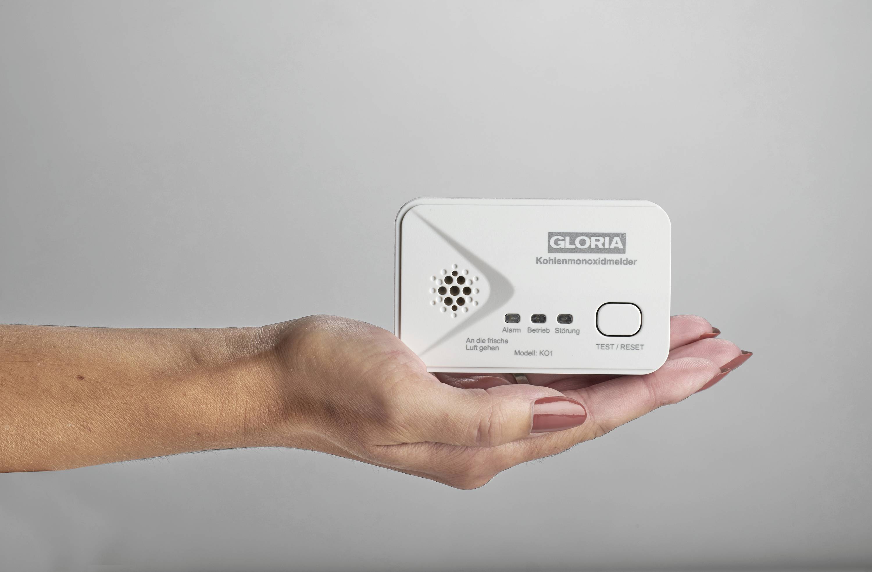 En hand håller en kolmonoxidvarnare märkt med 'GLORIA Kolmonoxidvarnare'. Varnaren har test- och återställningsknappar.