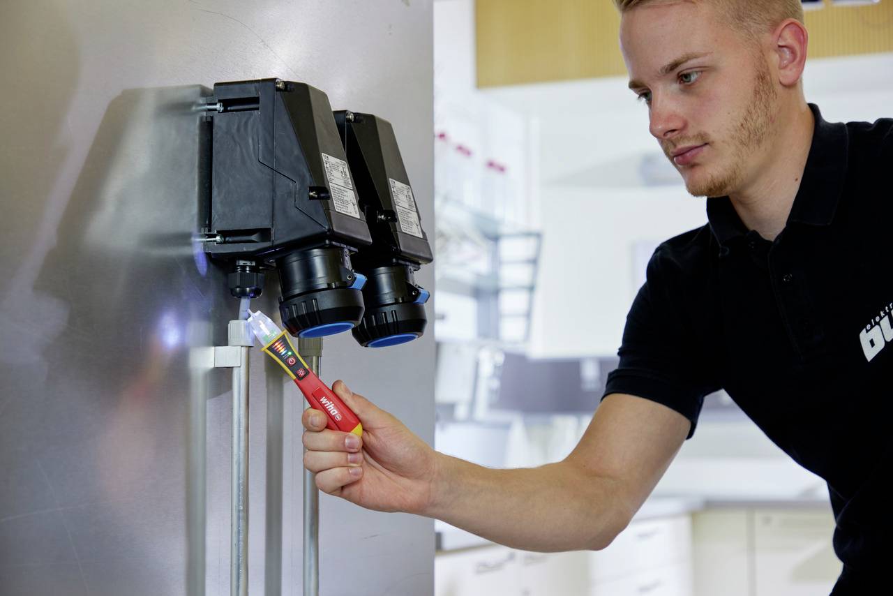 En tekniker i en svart polotröja kontrollerar maskinens elektriska system med en spänningstestare i en industriell verkstad.