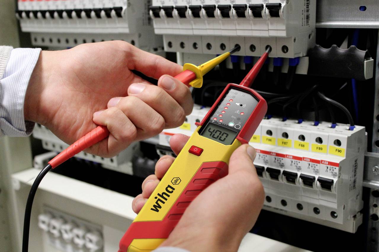 Två händer håller ett spänningsmätinstrument vid en öppen elskåp och mäter spänningen, som visas på displayen som '400V'.