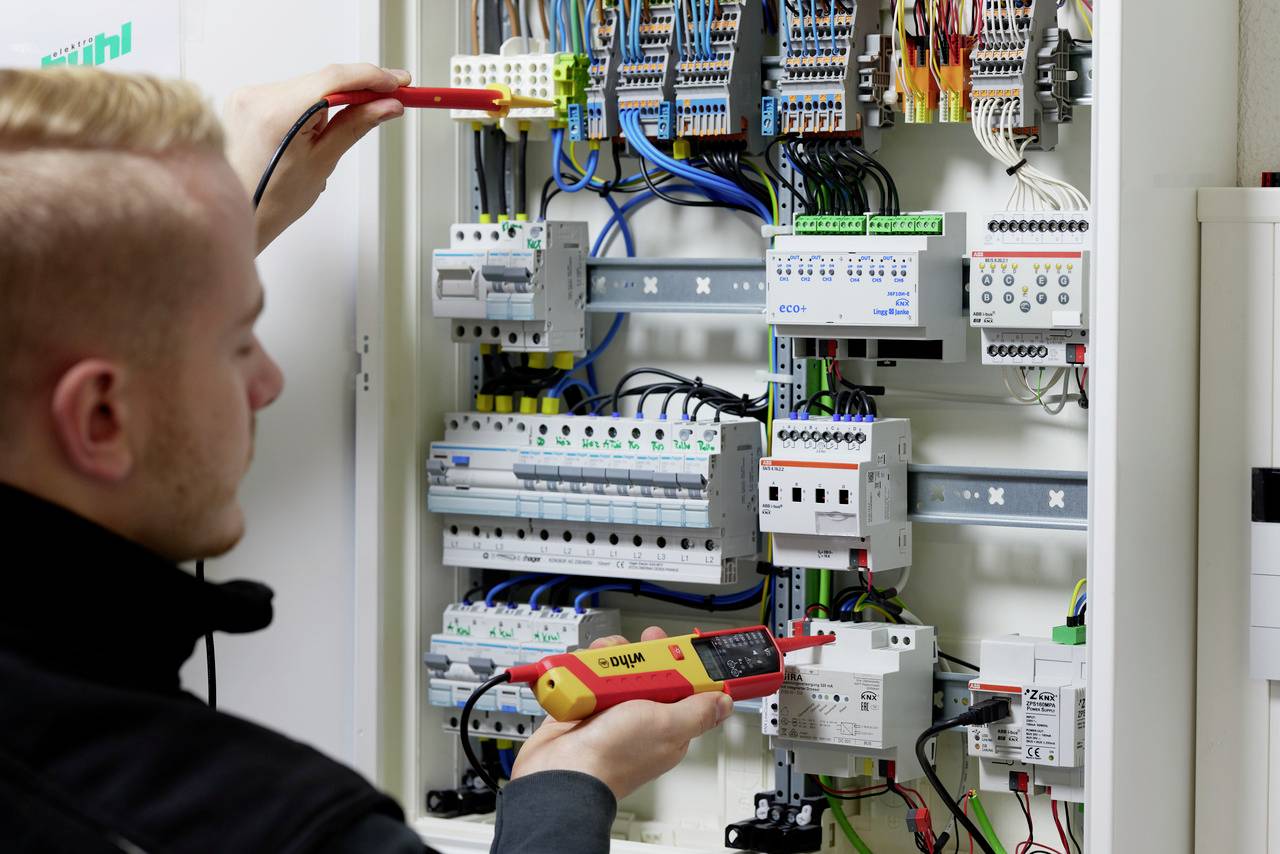 En tekniker kontrollerar kabeldragningen i en öppen säkringsbox med ett mätinstrument.