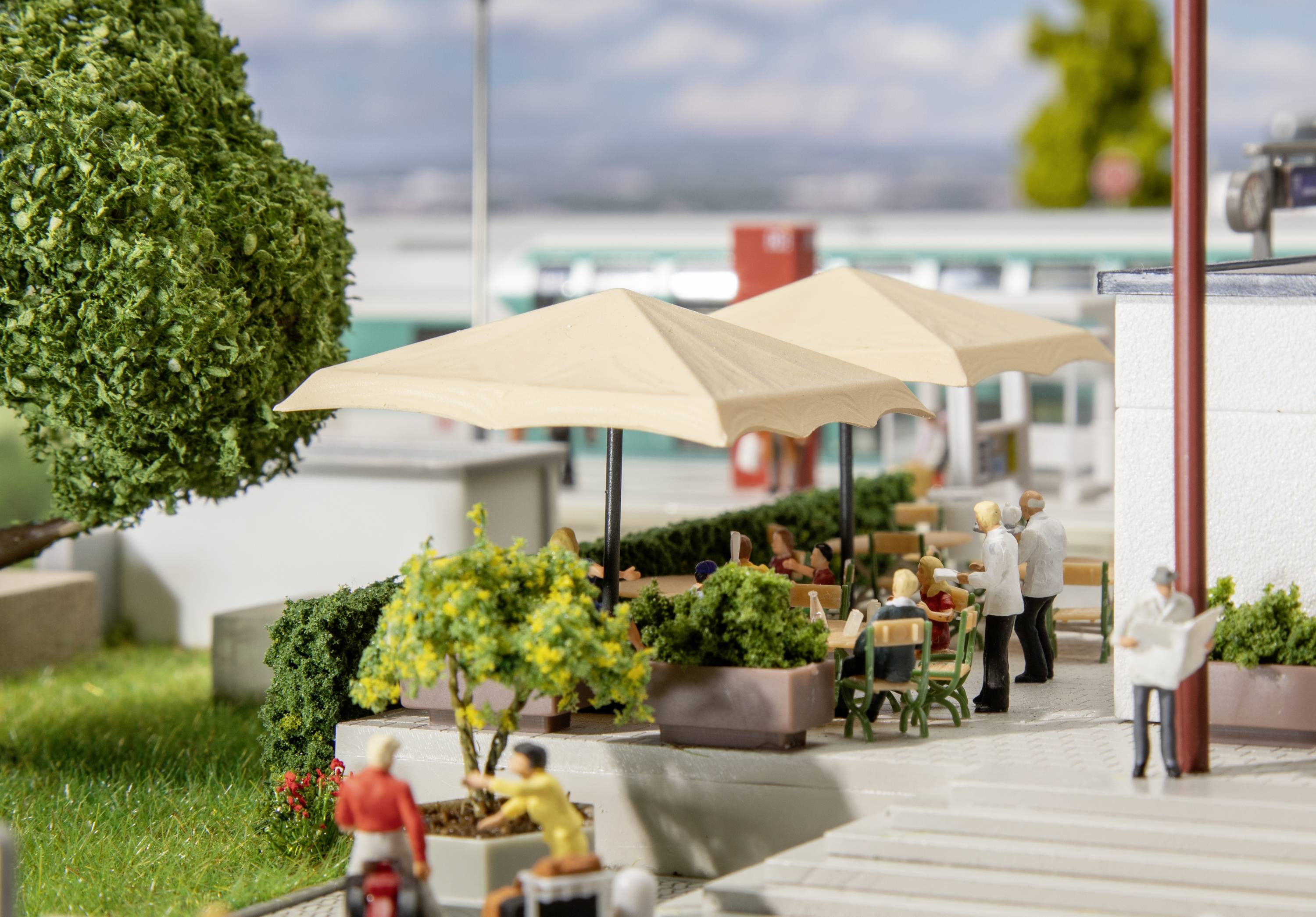 Miniatyrmodell av en gatuscen: Flera personer sitter under parasoll på ett uteserverat café, omgivna av växter.