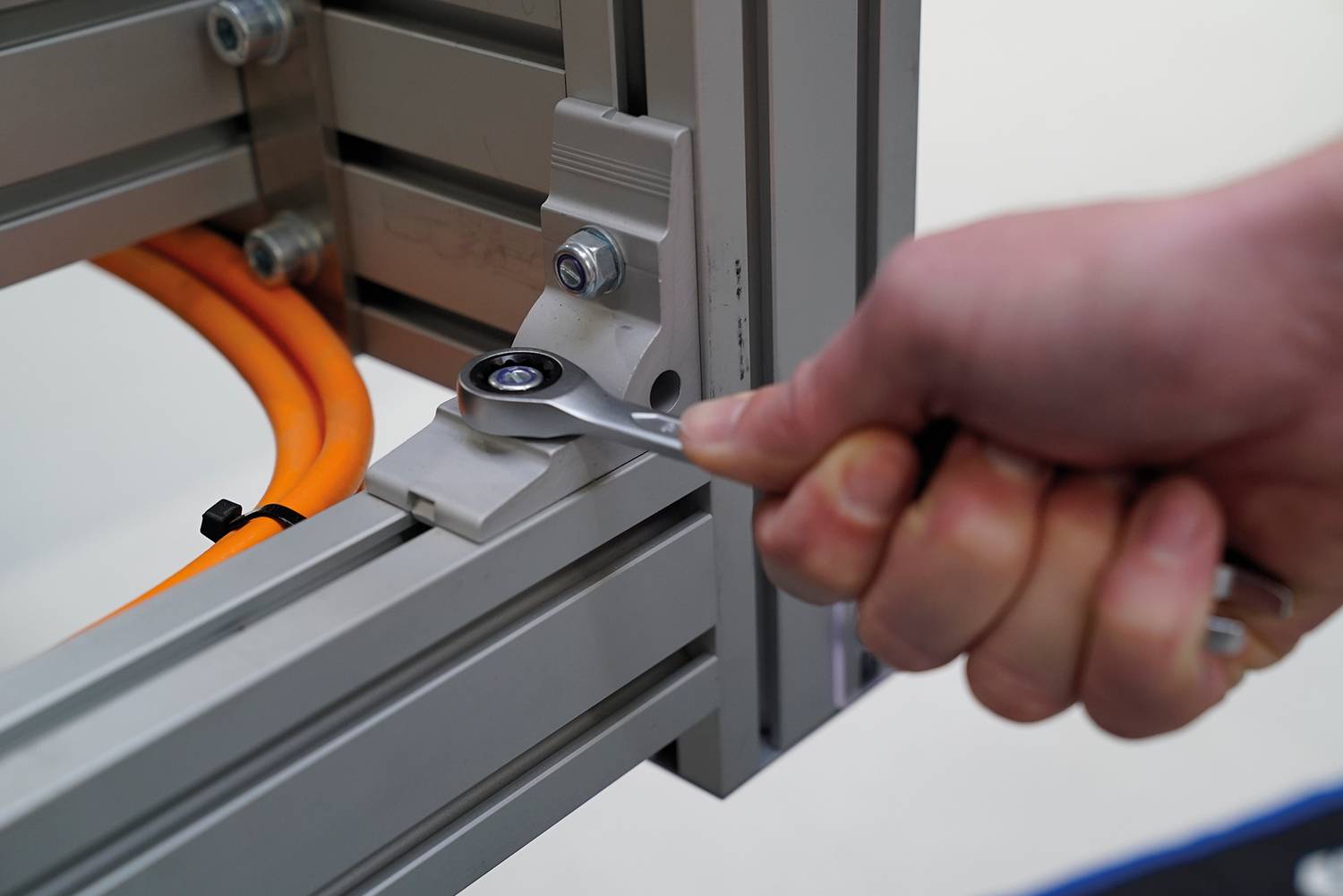En hand drar åt en skruv med en skiftnyckel på en metallisk maskinram med orange kabel.