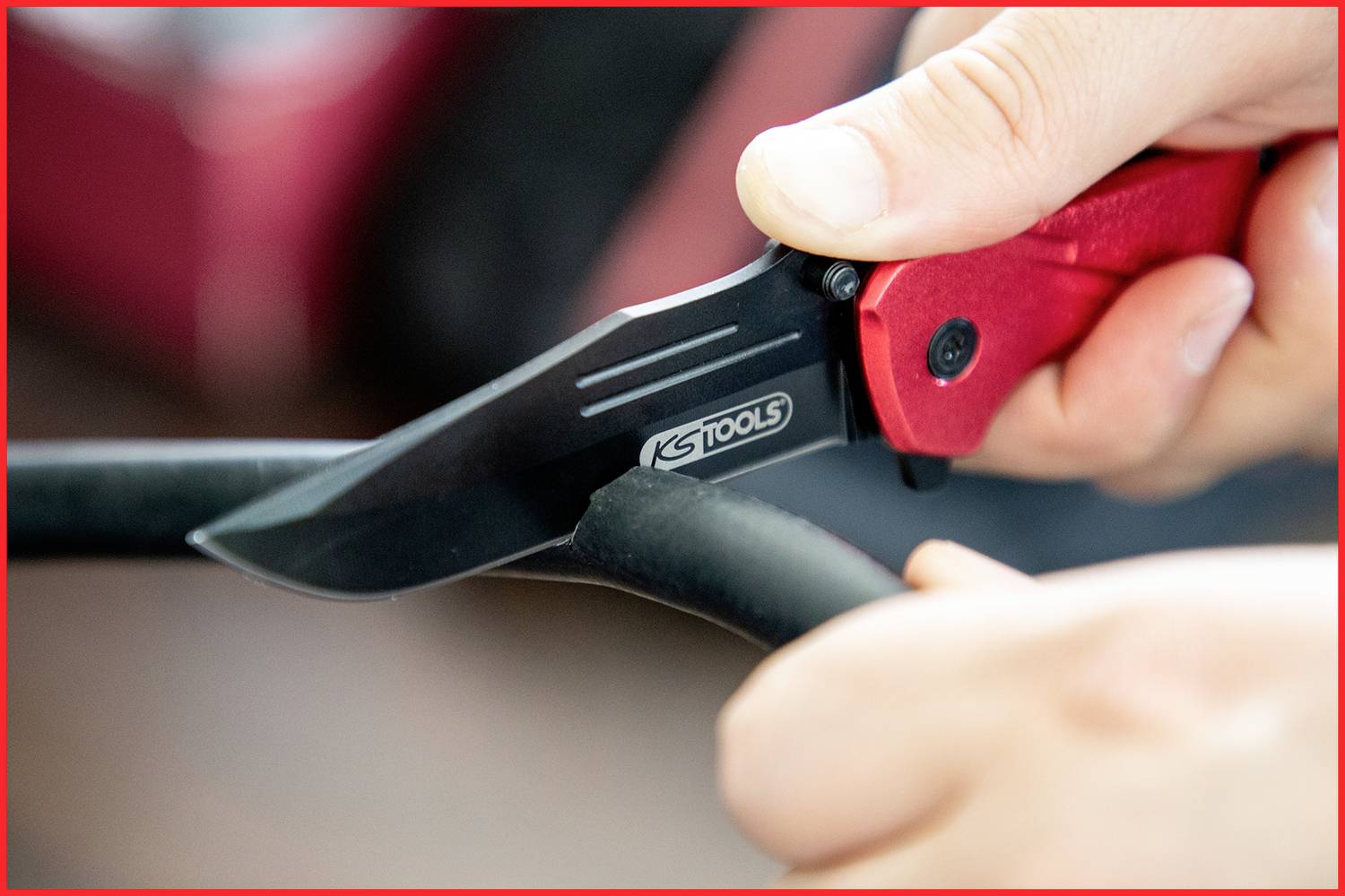 En hand håller en röd fällkniv och klipper genom en svart kabel eller slang. Kniven är märkt med 'KS TOOLS'.