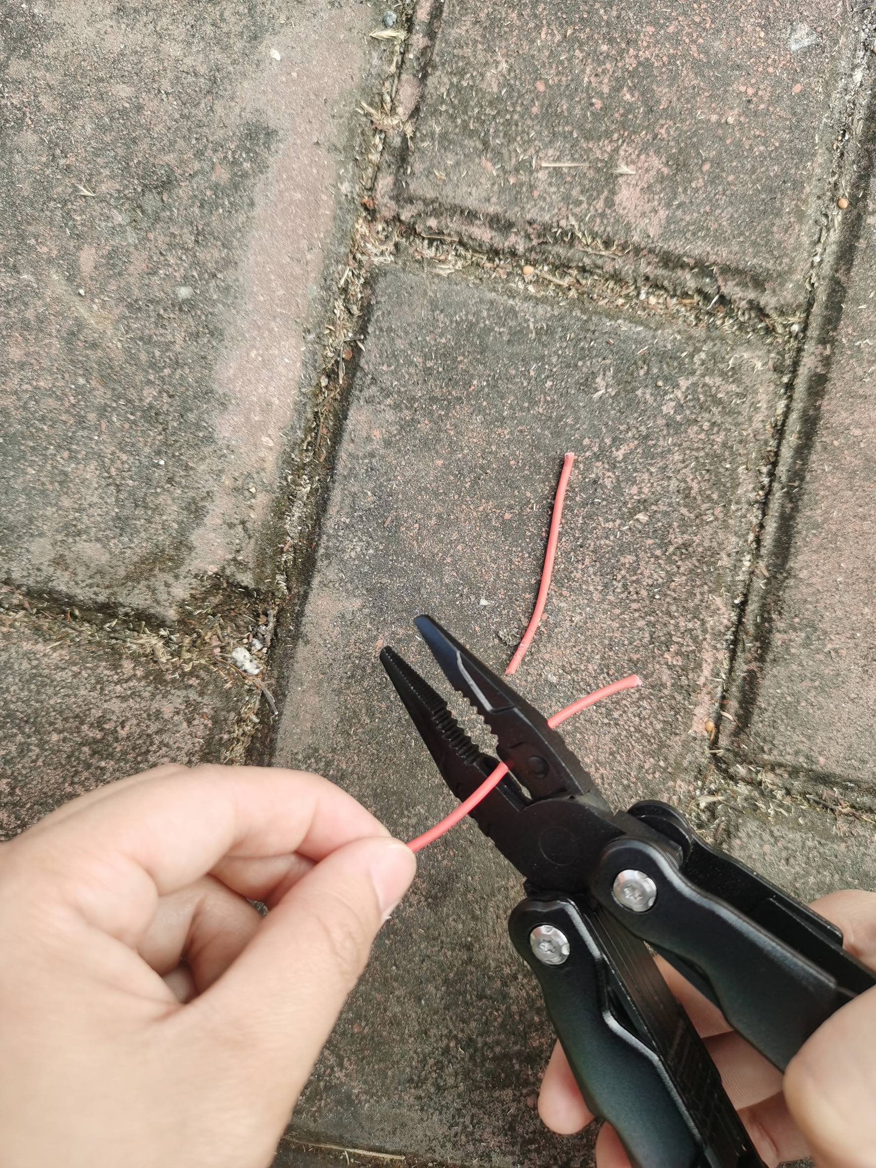 En hand håller en tång som klipper av en röd kabel. Bakgrunden består av gatsten.