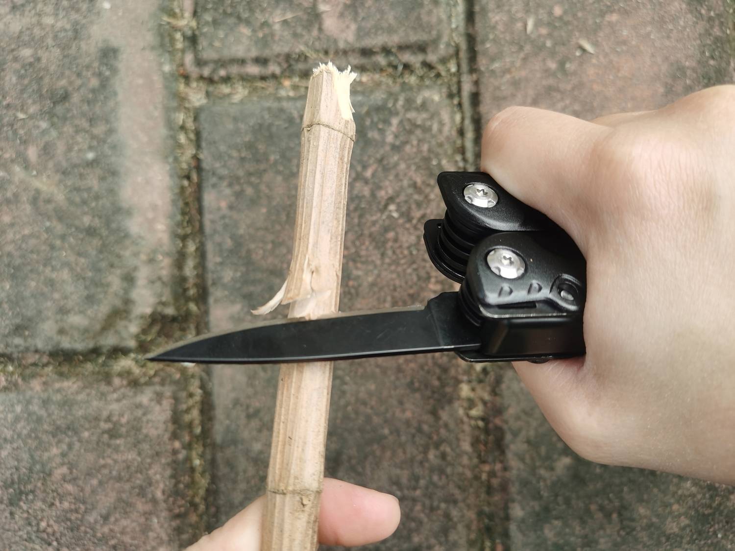 En hand snider med en svart fällkniv på en träpinne medan den står på ett stenlagt golv.