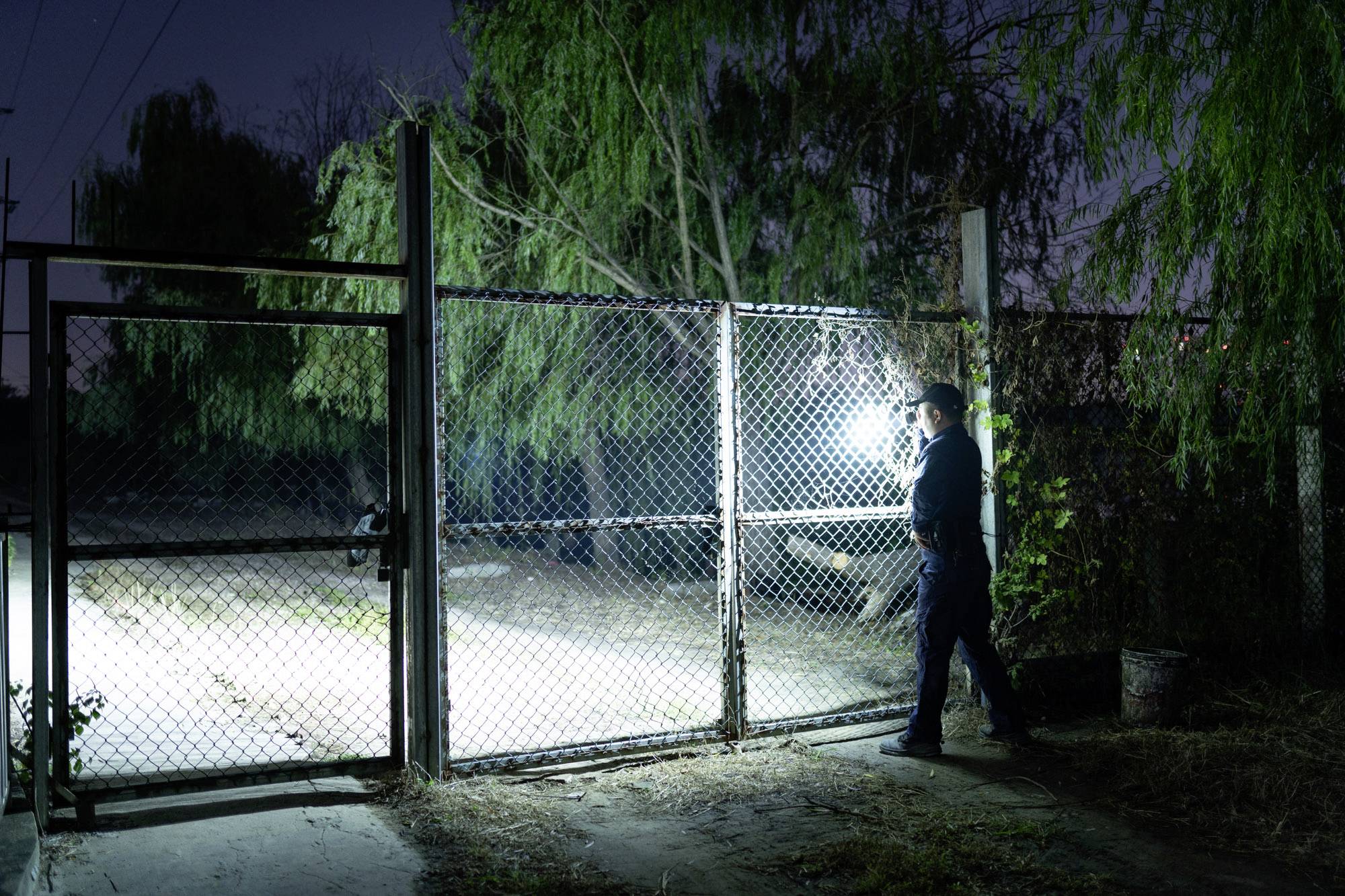 En person lyser med en ficklampa mot ett grindlås i en nattlig omgivning, medan träd syns i bakgrunden.