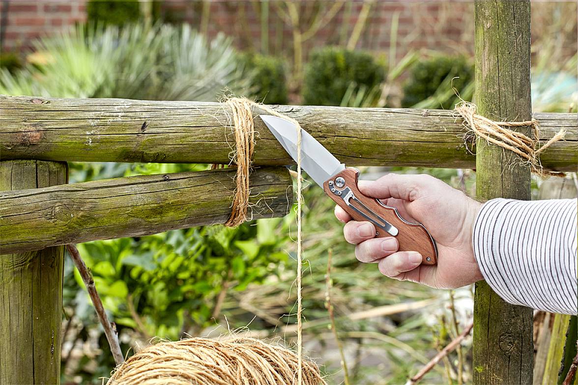 En person klipper sisal-band med en fällkniv från ett trästaket, troligen i en trädgård. I bakgrunden finns växter och buskar.