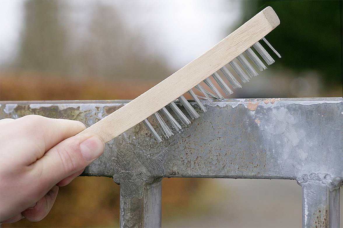En hand håller en ståltrådsborste som förs över en rostig metallyta för att avlägsna rost.