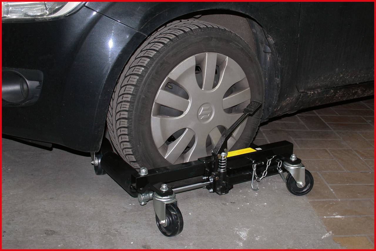 En svart bil står på ett parkeringsplan. En domkraft med hjul är placerad under det högra framhjulet.