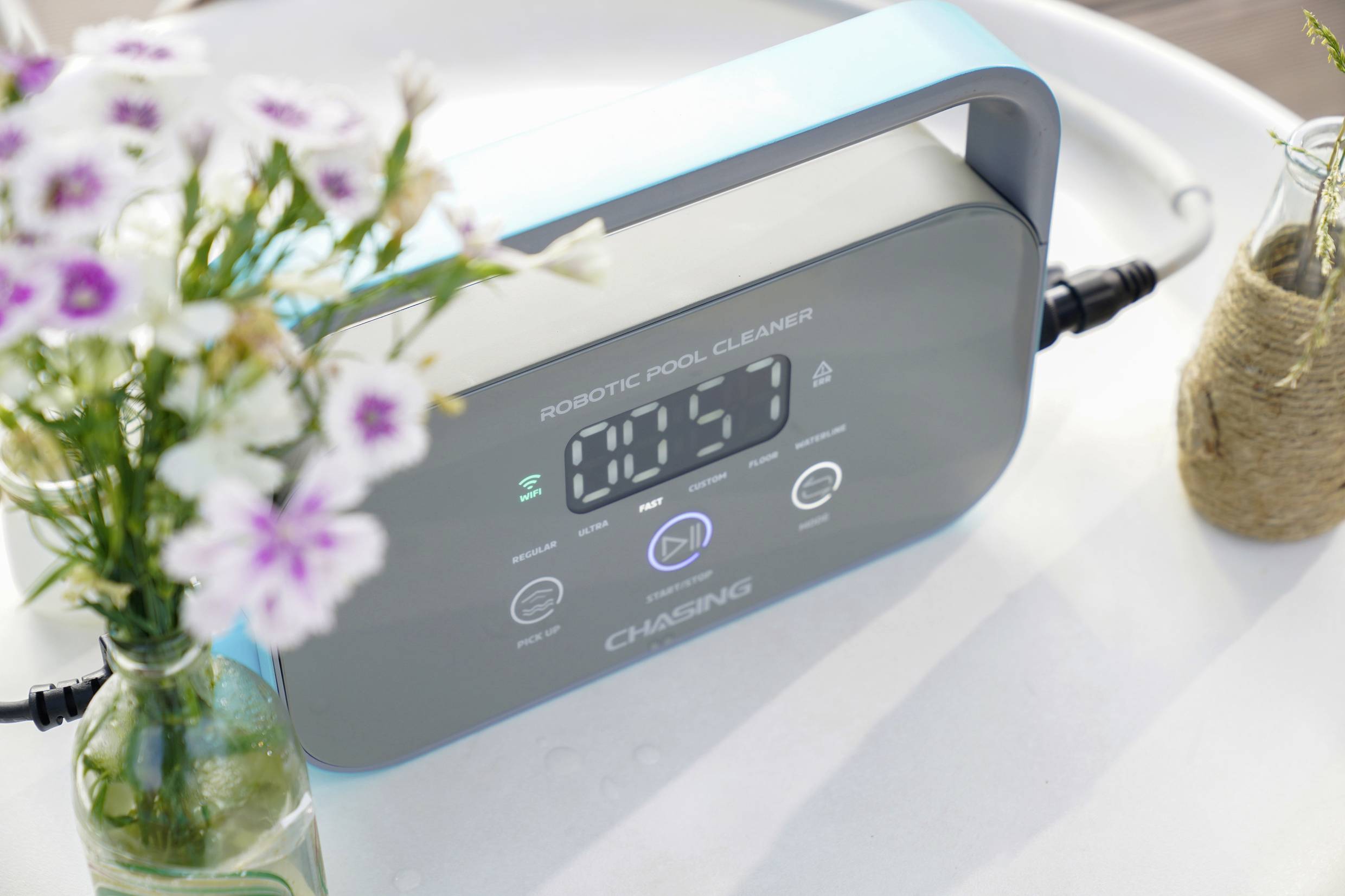 Digital display på en poolrobot med tiden '10:57', bredvid en vas med blommor. Enheten är från märket 'Chasing'.