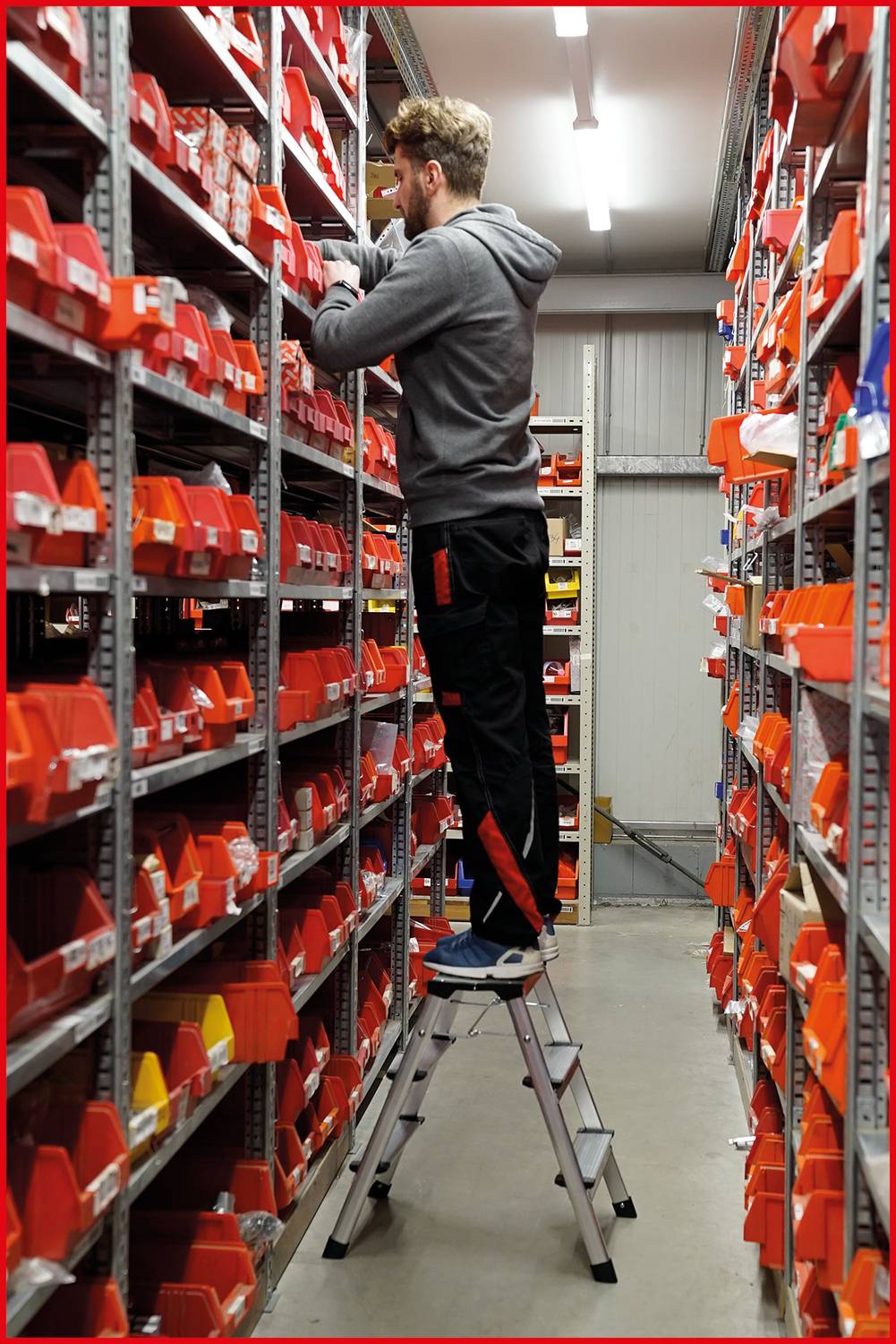 En person står på en stege i ett lager och organiserar röda plastbehållare i hyllorna.