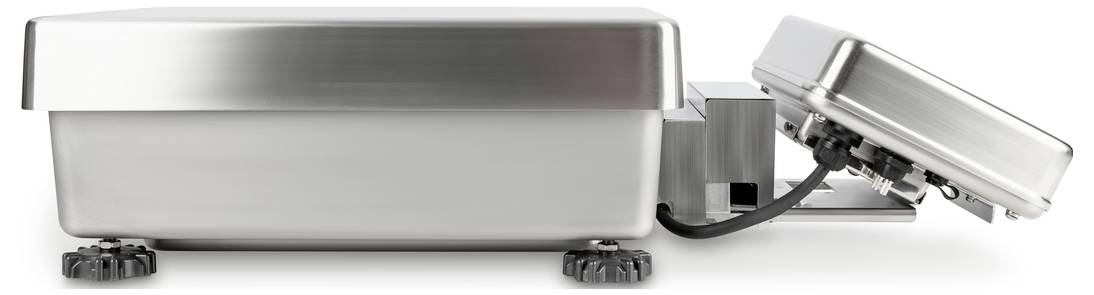 Rektangulär, metallisk låda med avtagbart lock, förbunden via en kabel. Locket ligger snett till höger mot underlaget.