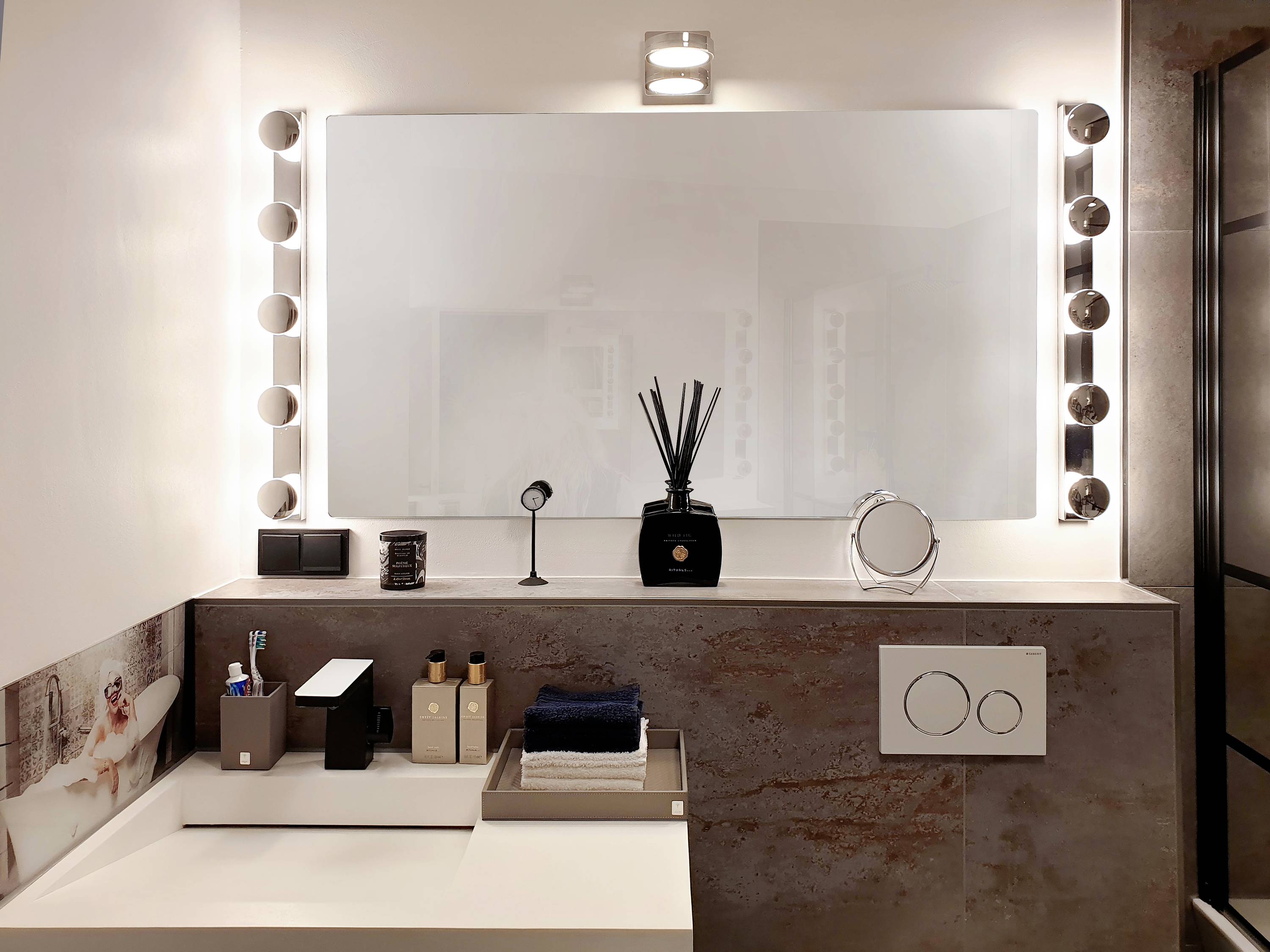 Modernt badrum med stor, belyst spegel, handfat med handdukar och hygienprodukter. En atmosfär av elegans och funktionalitet.