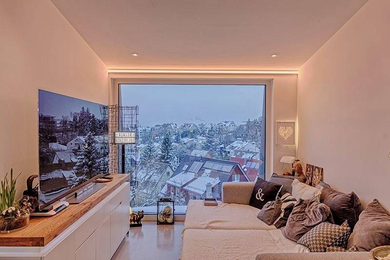 Vardagsrum med stora fönsterpartier, utsikt över snötäckta tak. Mysig soffa med många kuddar, modern TV till vänster.