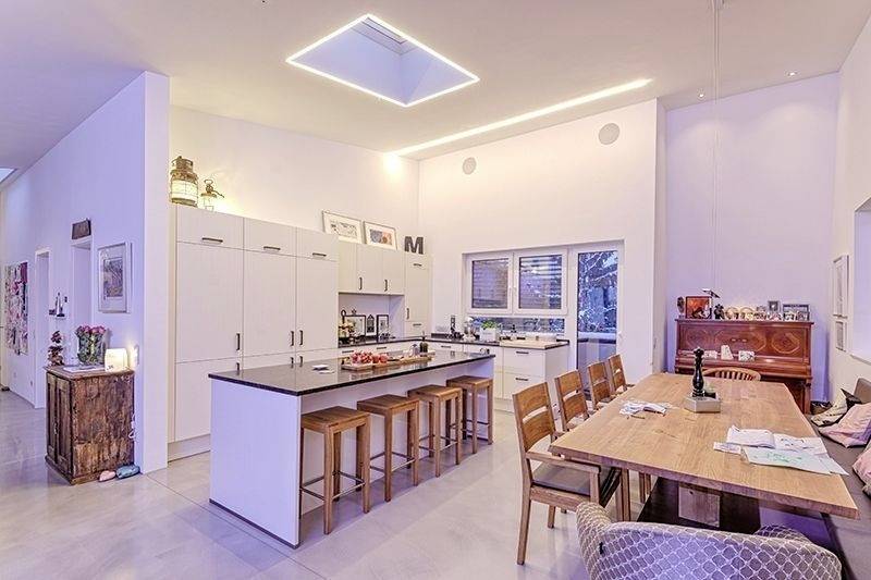 Modern kök och matområde med vitt design, central köksö med stolar, stort matbord, piano och diskreta takarmaturer.